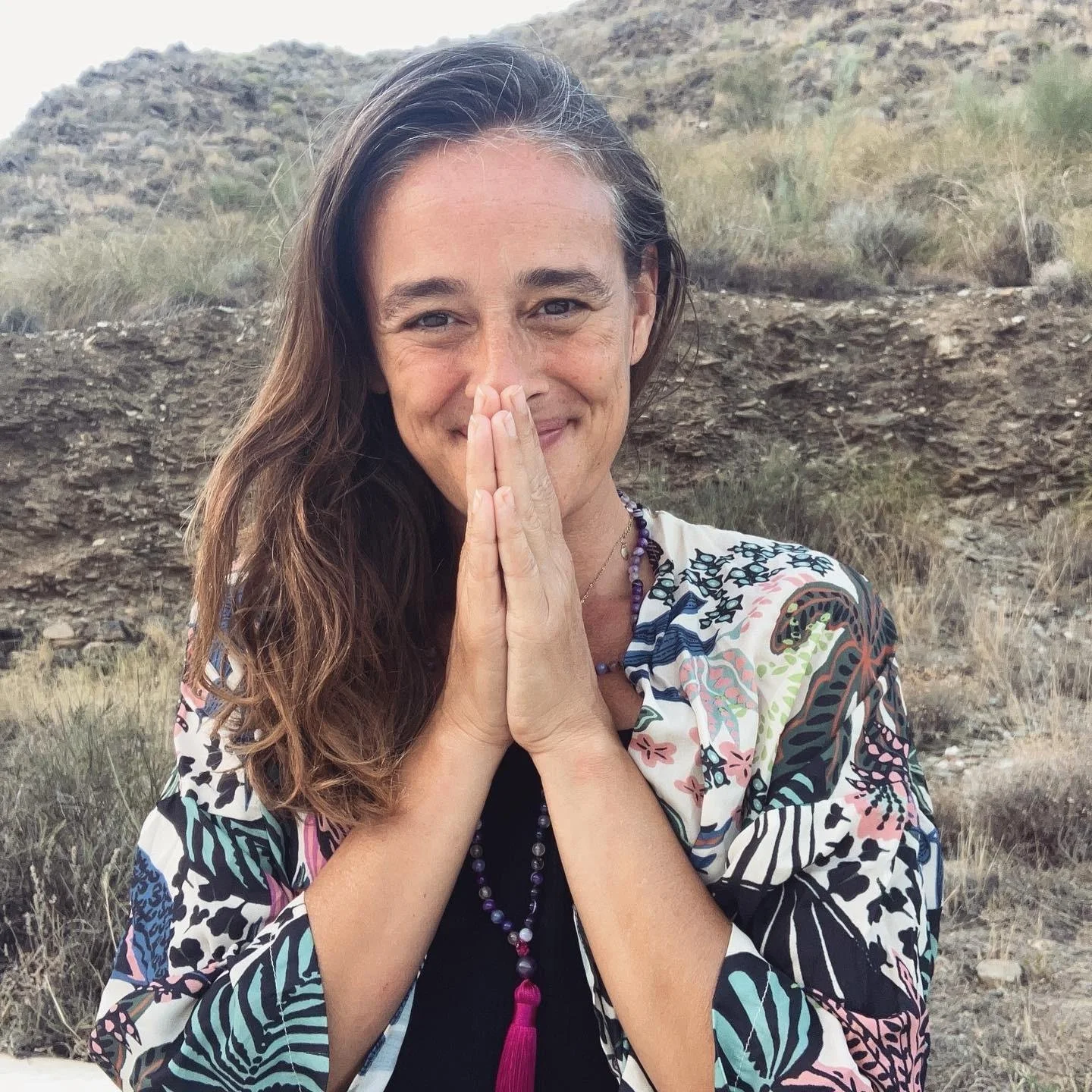 A woman with long, wavy brown hair smiling and holding her hands together near her face in a prayer-like gesture outdoors in a dry, rocky landscape with sparse vegetation.