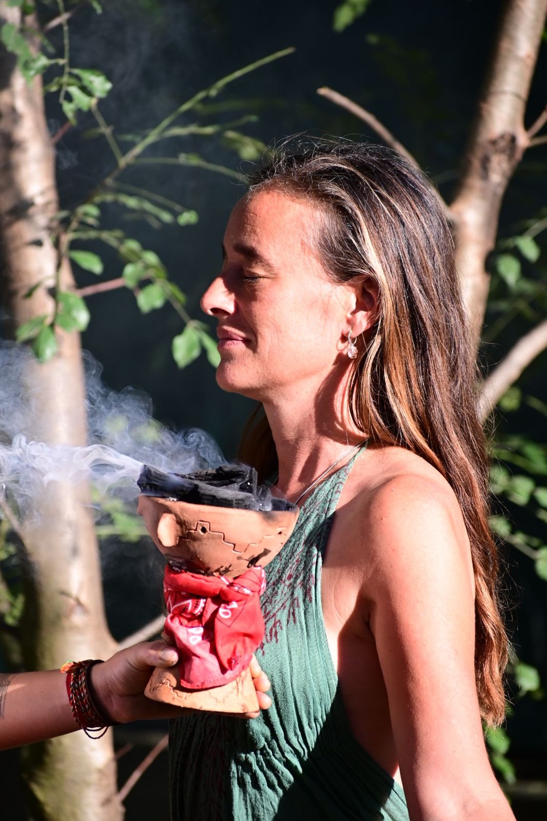 A woman with long brown hair and an earring, wearing a green sleeveless top, is standing outdoors near trees, holding a burning incense holder with smoke rising, and smiling with her eyes closed.