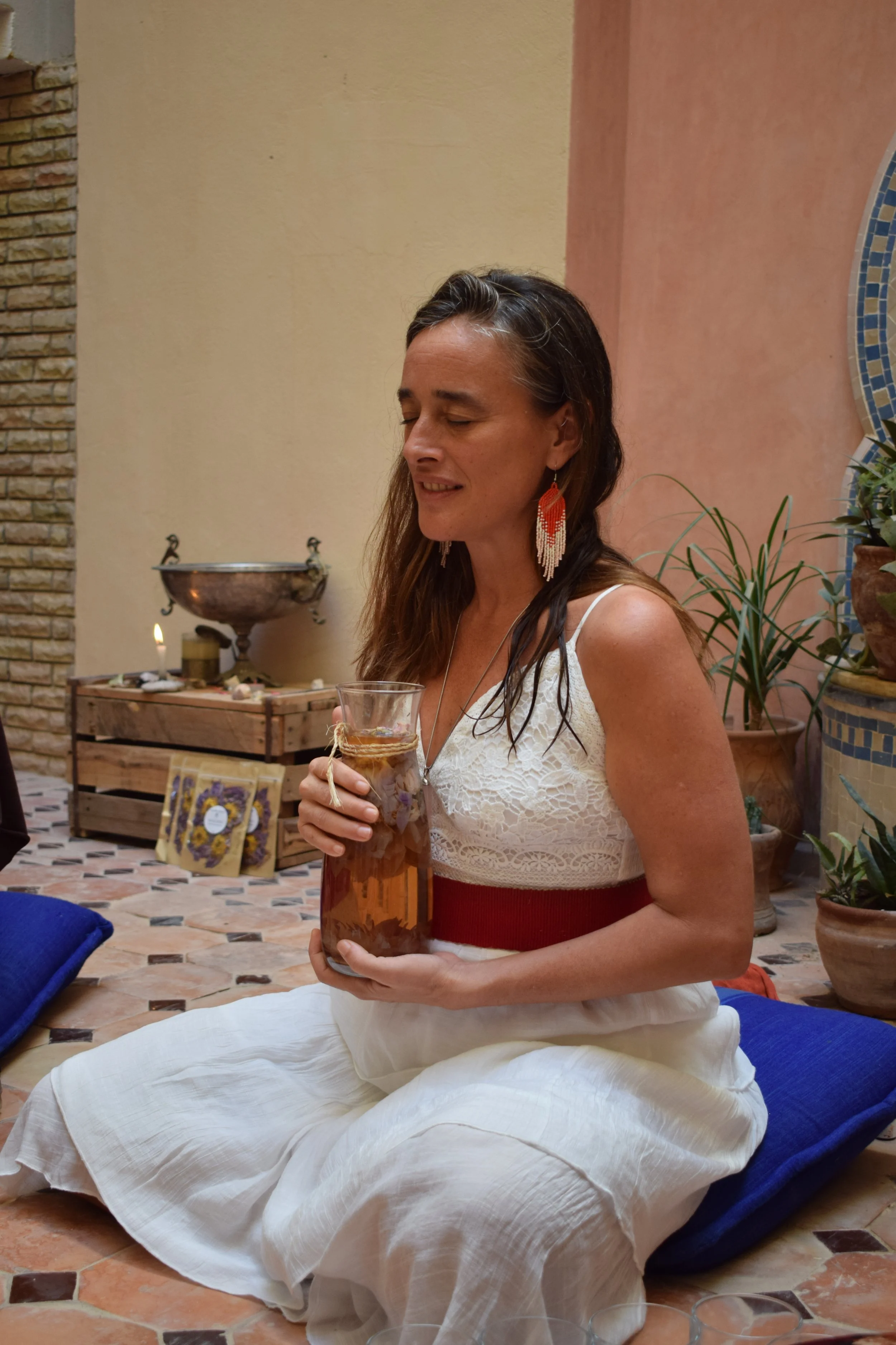 A woman with long dark hair, wearing earrings and a white lace top, is sitting cross-legged on the floor holding a glass of tea. She appears to be in a serene or meditative state, with her eyes closed, in a warmly decorated room with potted plants an