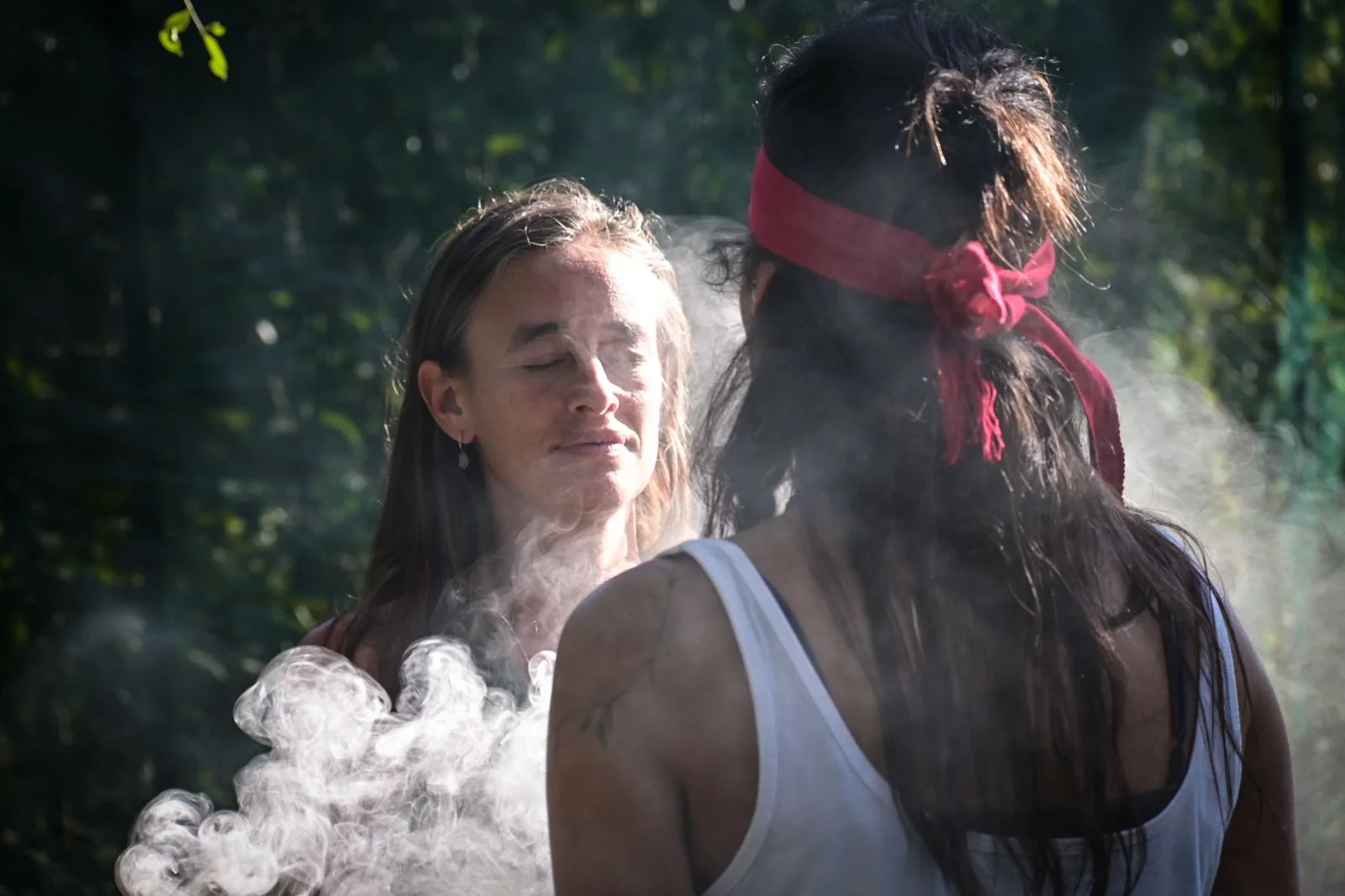 Two women face each other outdoors with steam or smoke around them. One woman has long dark hair, a red headband, and a white tank top. The other woman has light brown hair and wears a white top. The scene is set in a wooded or garden area with trees