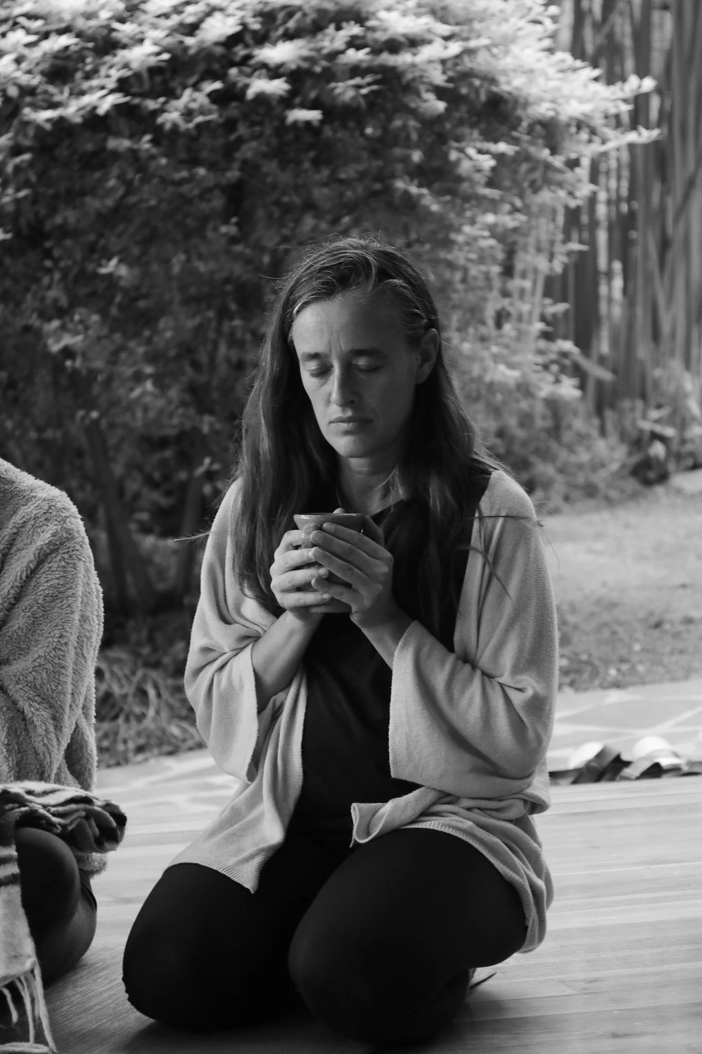 A woman with long hair, sitting cross-legged on the floor outdoors, looking at her phone with her eyes closed. She is wearing a light jacket and dark clothing, and there are trees and a wooden fence in the background.