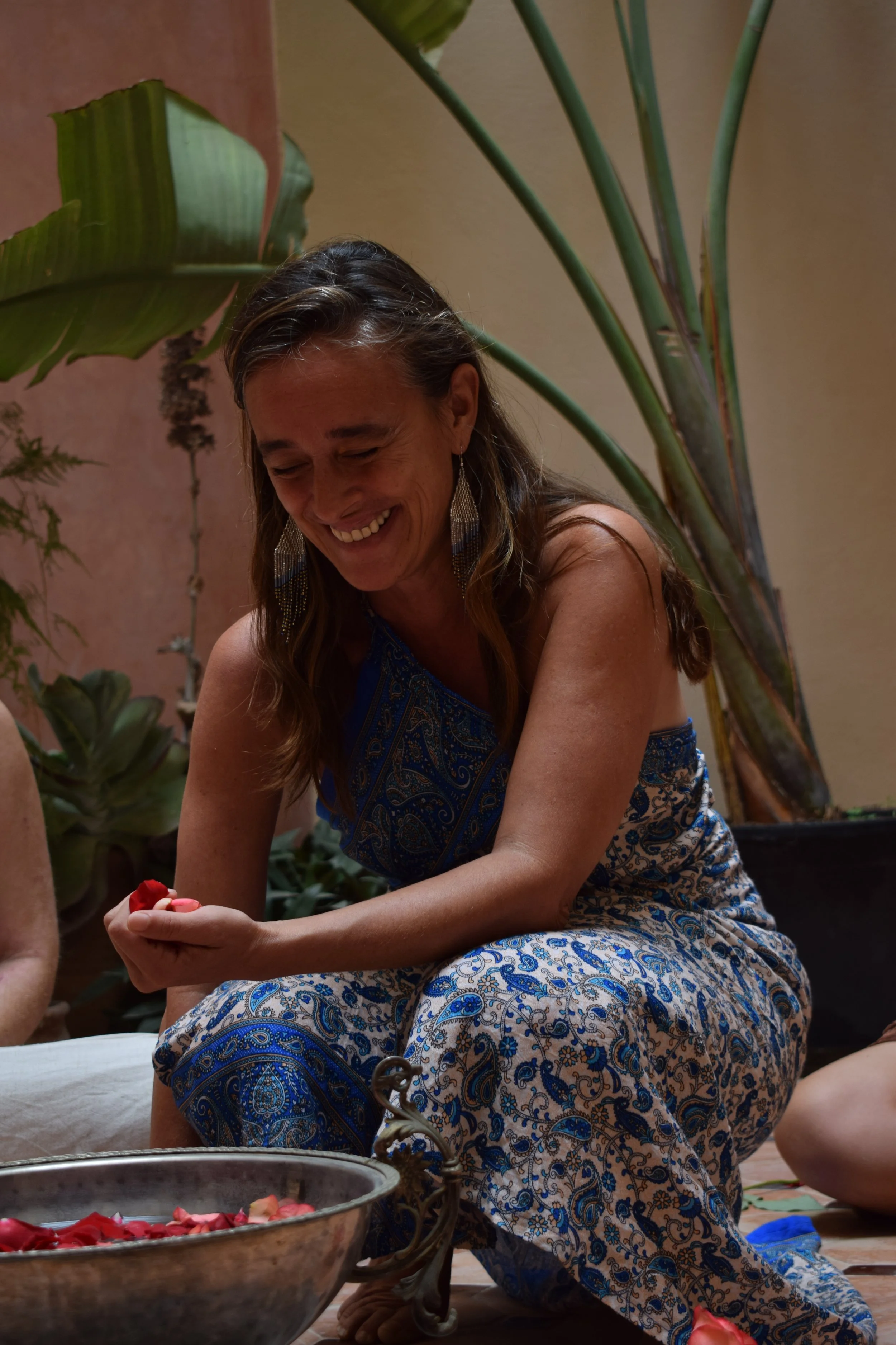 A woman smiling and sitting cross-legged on the floor, surrounded by large green indoor plants, wearing a blue patterned dress and large earrings.