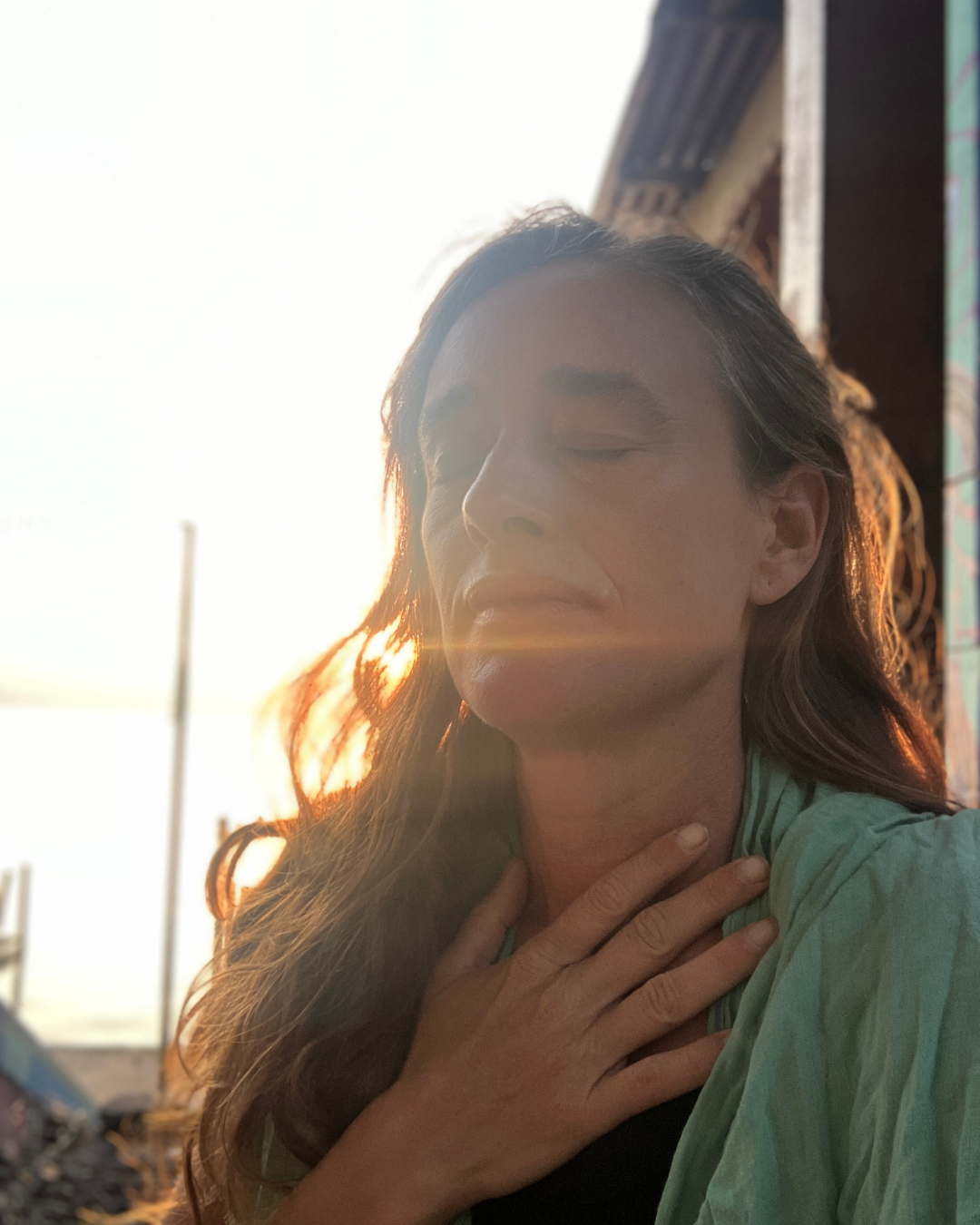 A woman with long hair, eyes closed, holding her hand to her neck, standing outdoors during sunset with sunlight illuminating her face.