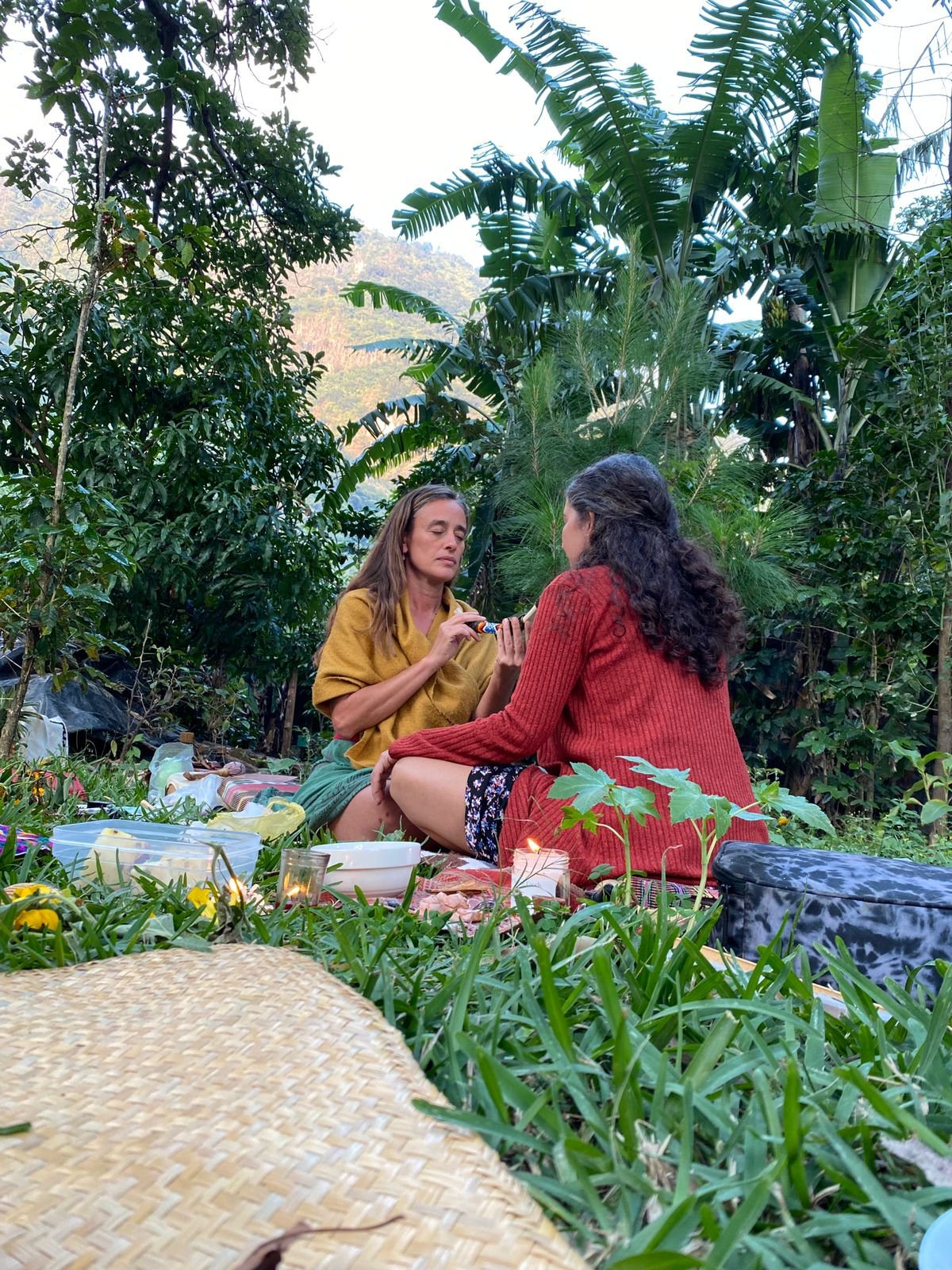 Two women sitting on a blanket in a lush garden with tropical plants, having a tea or picnic, surrounded by food, drinks, and candles, with mountains in the background.