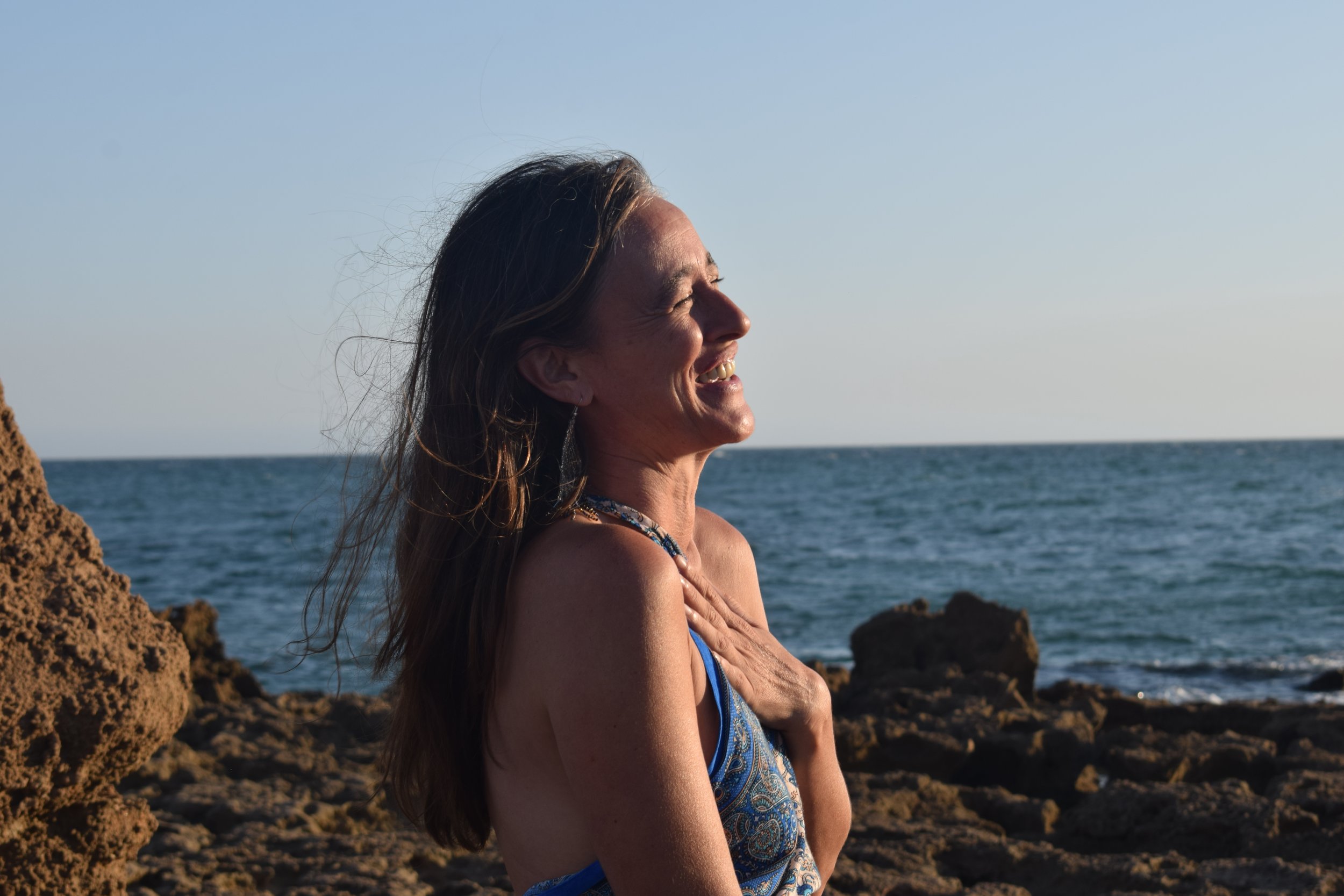 A woman with long hair smiling and looking to the right at the seaside with rocks and the ocean in the background.