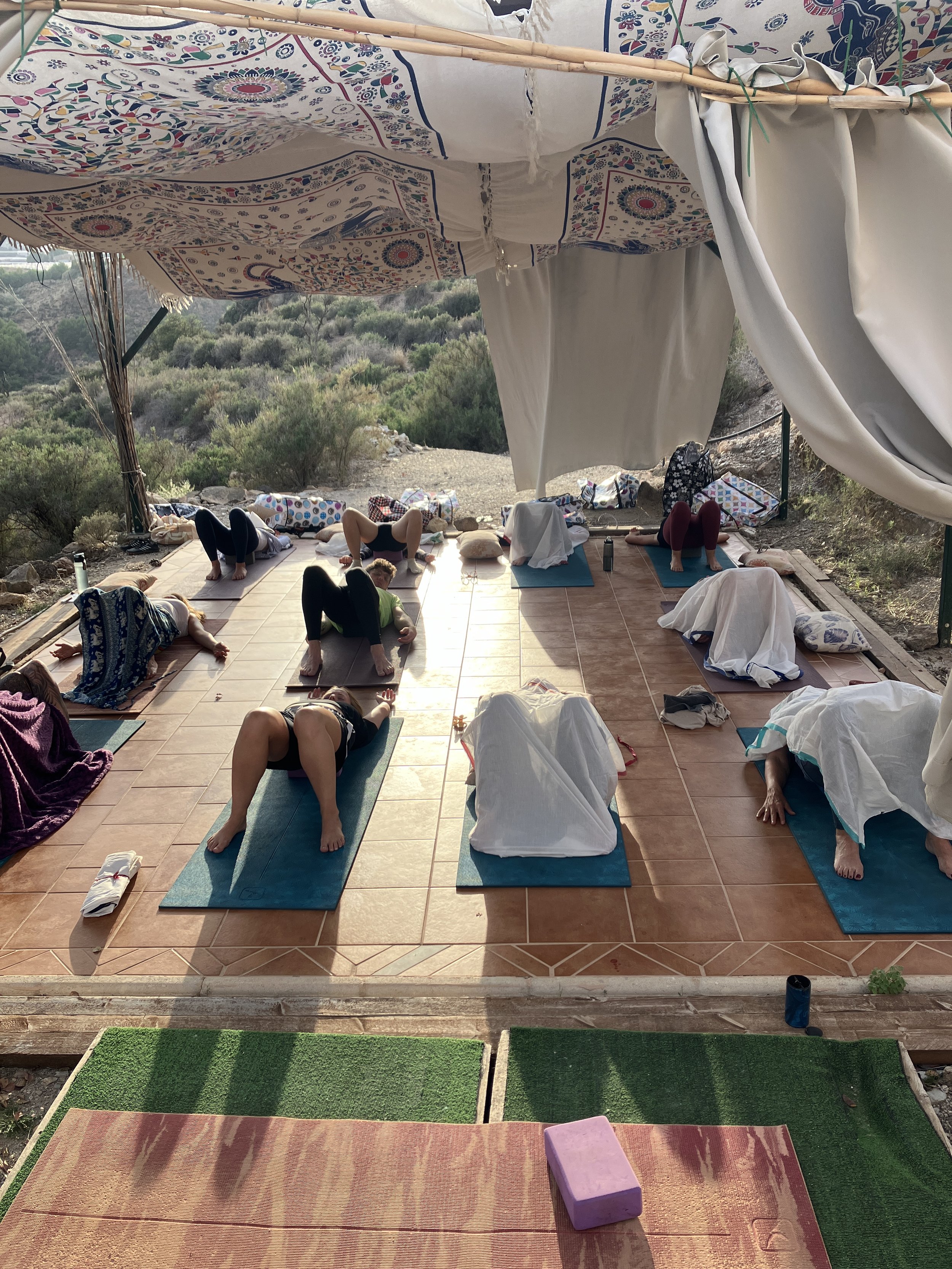 A group of people practicing yoga on mats outdoors on a tiled patio, with some covered by blankets or towels, surrounded by natural landscape with bushes and hills, under a canopy with colorful patterned fabric.