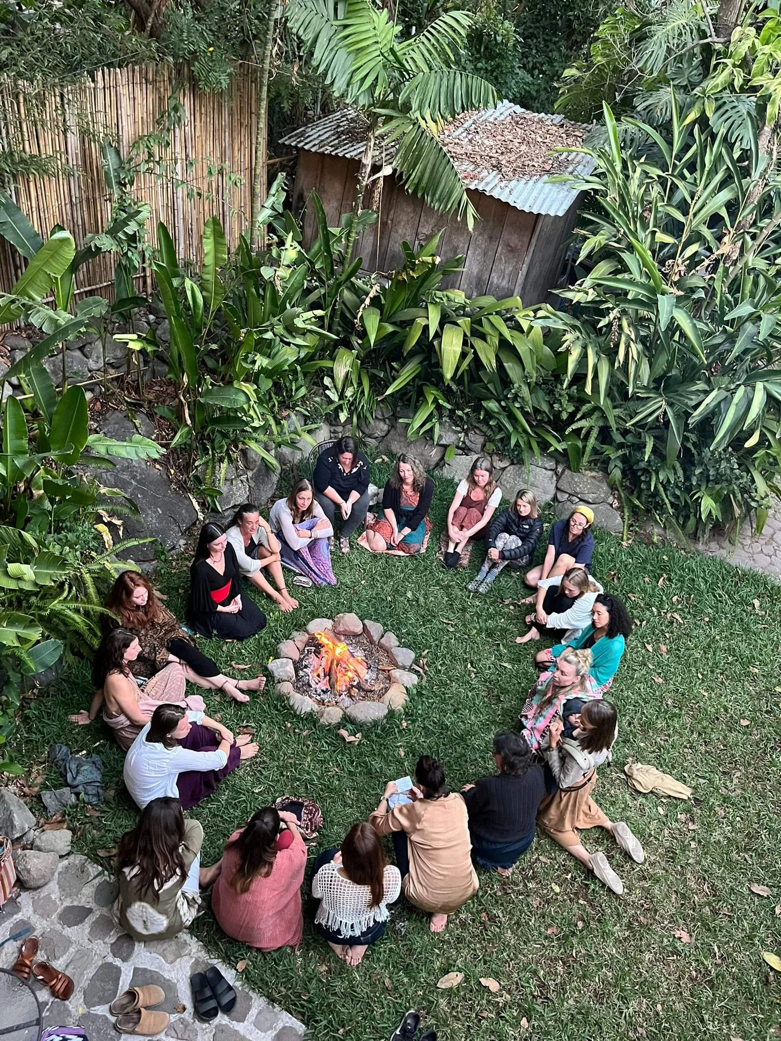 Group of people sitting in a circle around a small fire in a lush, green outdoor setting. Some individuals are sitting on rocks or grass, others on blankets. The area is surrounded by tall plants and trees, creating a serene atmosphere.