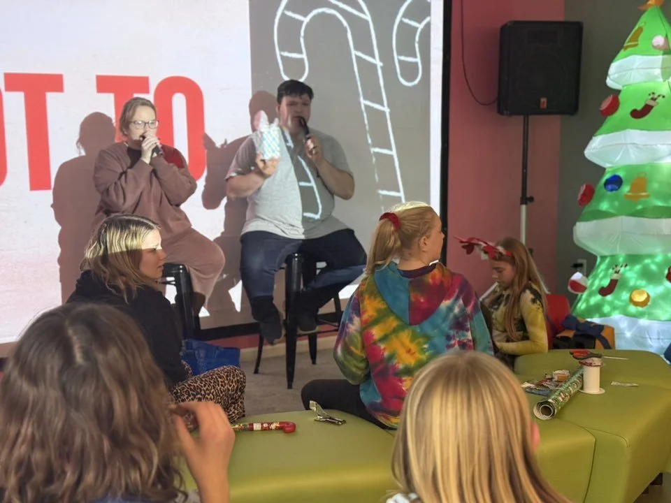 Two people sitting on stools with microphones on stage, one holding a gift bag, in front of a Christmas decoration with an inflatable Christmas tree, and a group of children sitting and standing nearby.