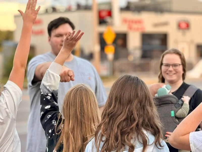 Group of people outdoors, some raising their hands, one person in the background giving a thumbs-up, smiling woman holding a baby in a carrier, blurry background of buildings and traffic.