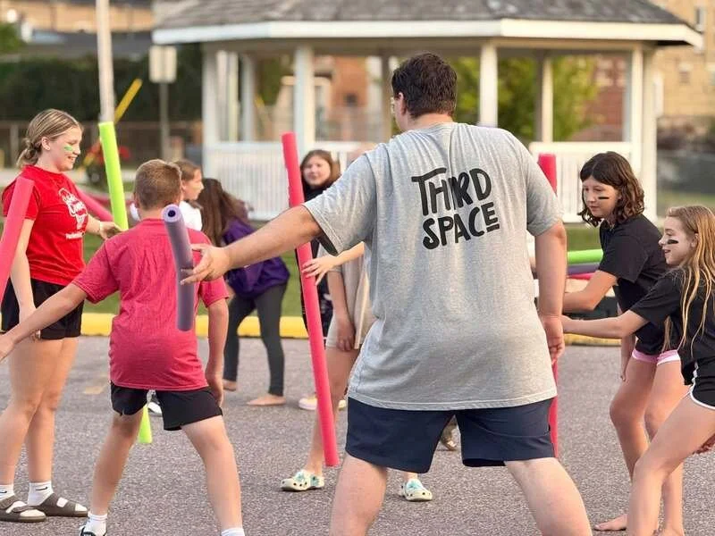 Children playing a game with foam pool noodles in an outdoor park area, supervised by an adult wearing a gray T-shirt that says 'Third Space'.