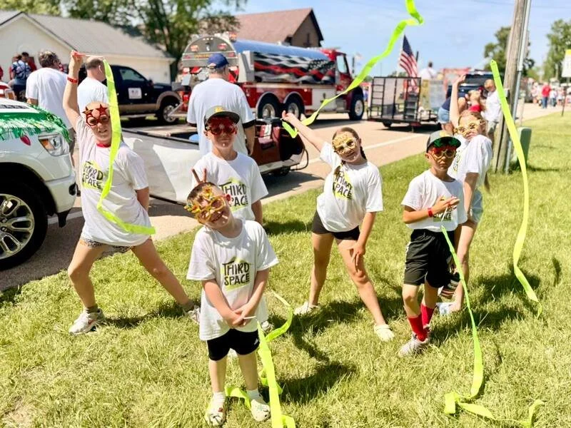 Children wearing masks and T-shirts that say 'Third Space,' holding yellow streamers and celebrating outdoors near parked cars and a fire truck during daytime.