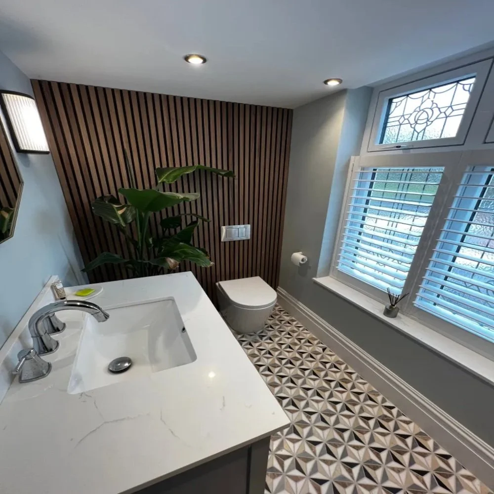 Modern downstairs toilet room in Allerton featuring a grey vanity unit with white basin, chrome tap, geometric patterned floor tiles, wood slat feature wall, wall-hung toilet, large window with shutters, and indoor plant for styling
