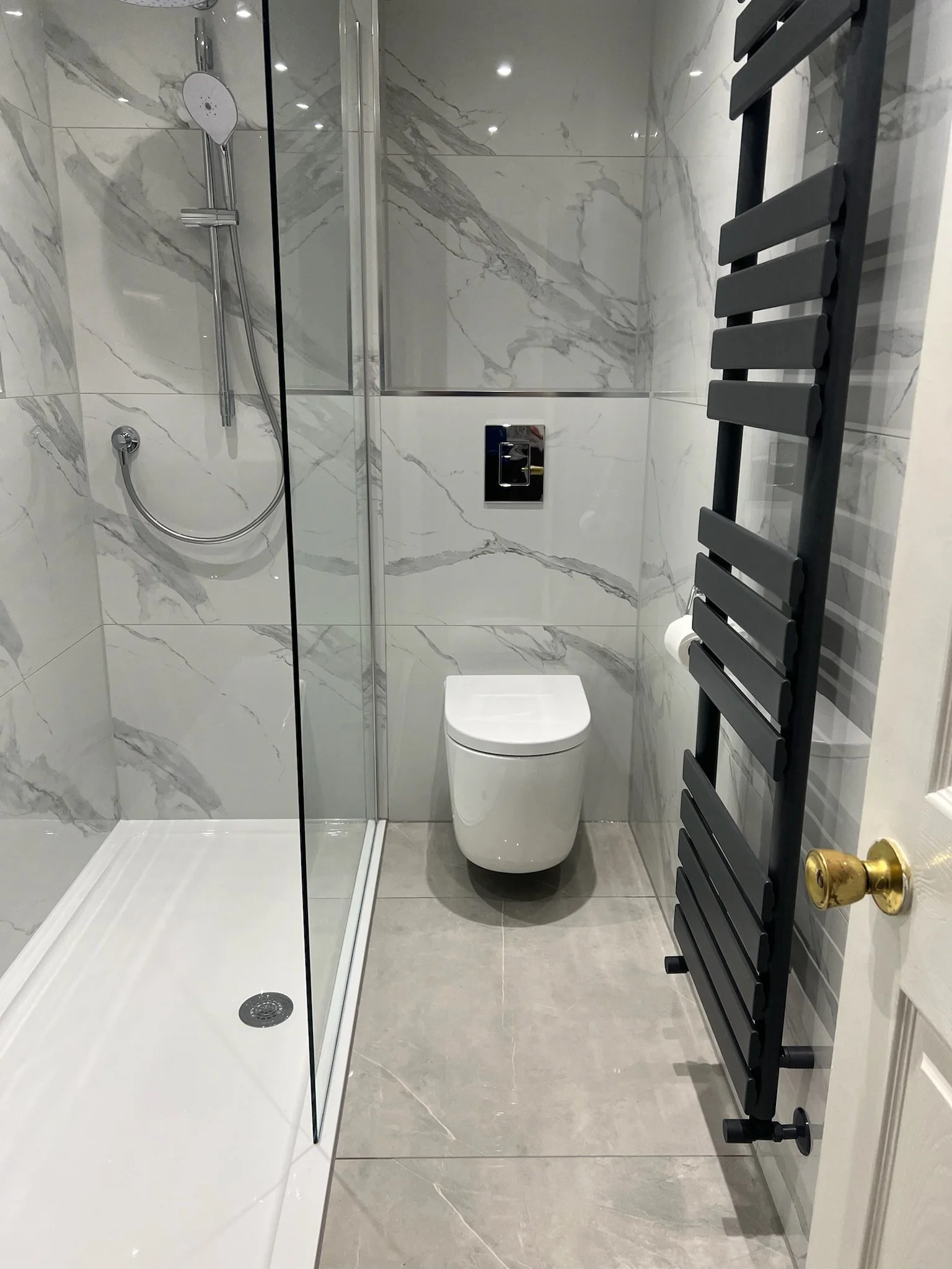 A sleek and modern bathroom renovation in Merseyside featuring a walk-in glass shower with elegant marble-effect wall panels and a minimalist wall-hung toilet. The space is complemented by neutral floor tiles and a tall black heated towel rail, creat