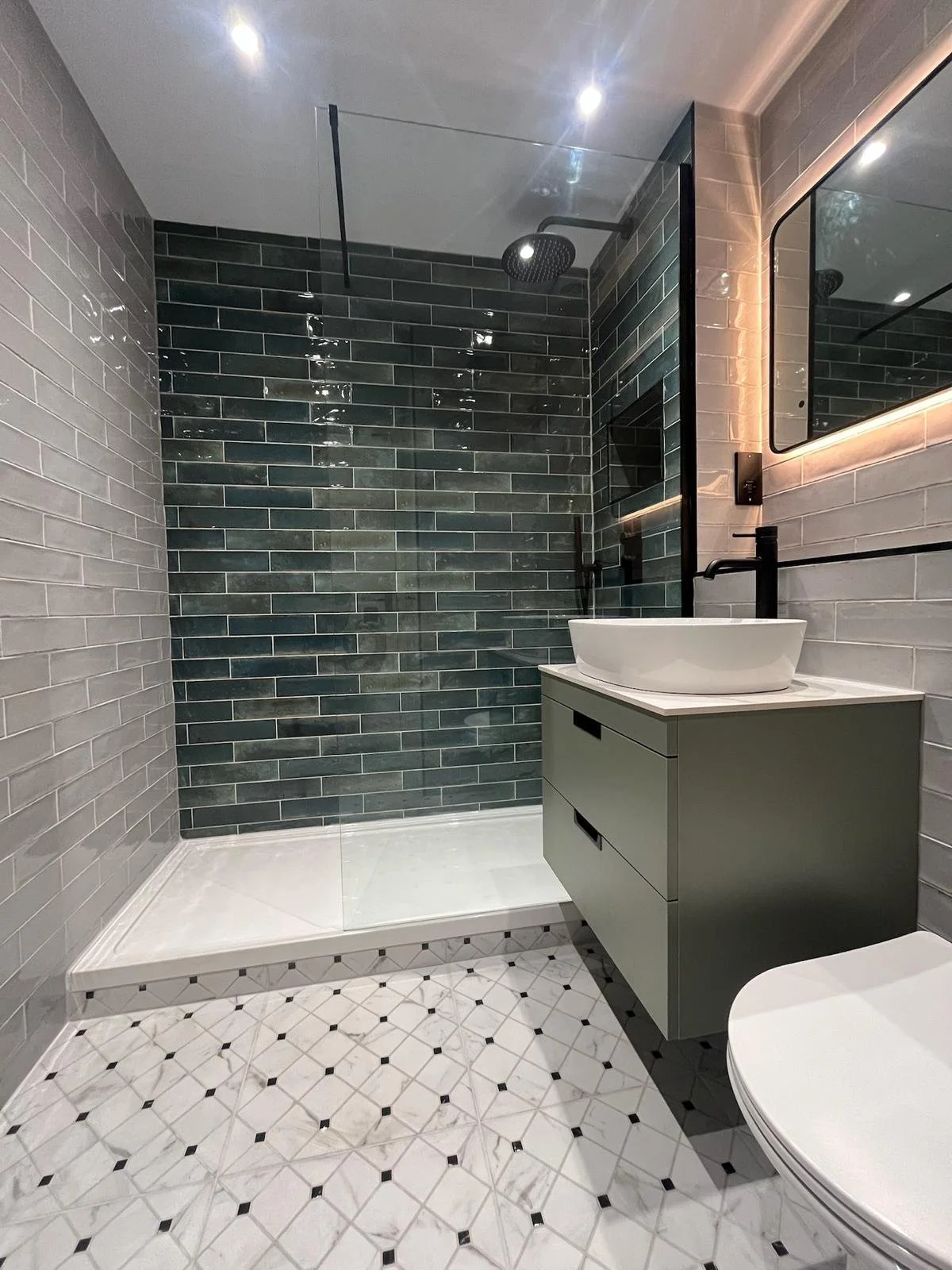 A stylish bathroom renovation in Merseyside featuring a walk-in shower with deep green brick-effect tiles and a sleek glass screen. The space includes a modern vanity unit with a countertop basin, a softly illuminated mirror, and classic patterned fl