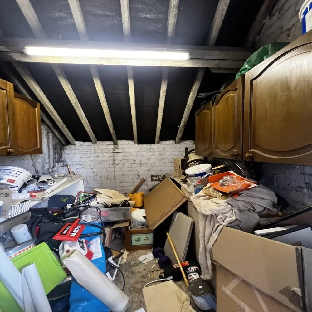 Overfilled loft space with exposed rafters, old wall cabinets, tools, and building materials awaiting clearance before bathroom renovation work in Bebington