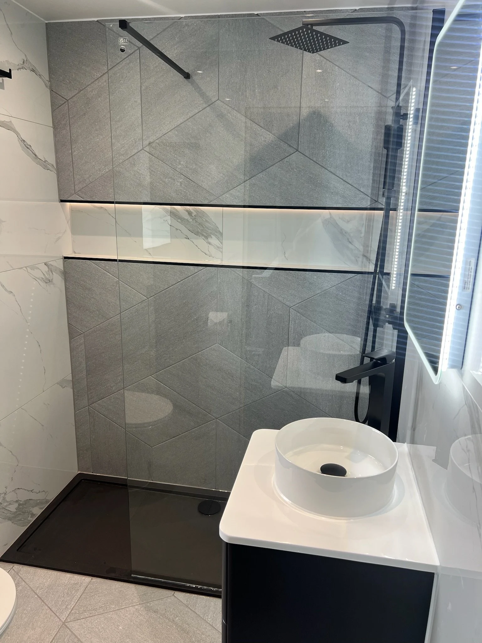 A contemporary bathroom renovation in Merseyside featuring a sleek walk-in shower with geometric grey wall tiles and a rainfall showerhead. The space includes a compact vanity unit with a countertop basin and matte black fittings, complemented by mar