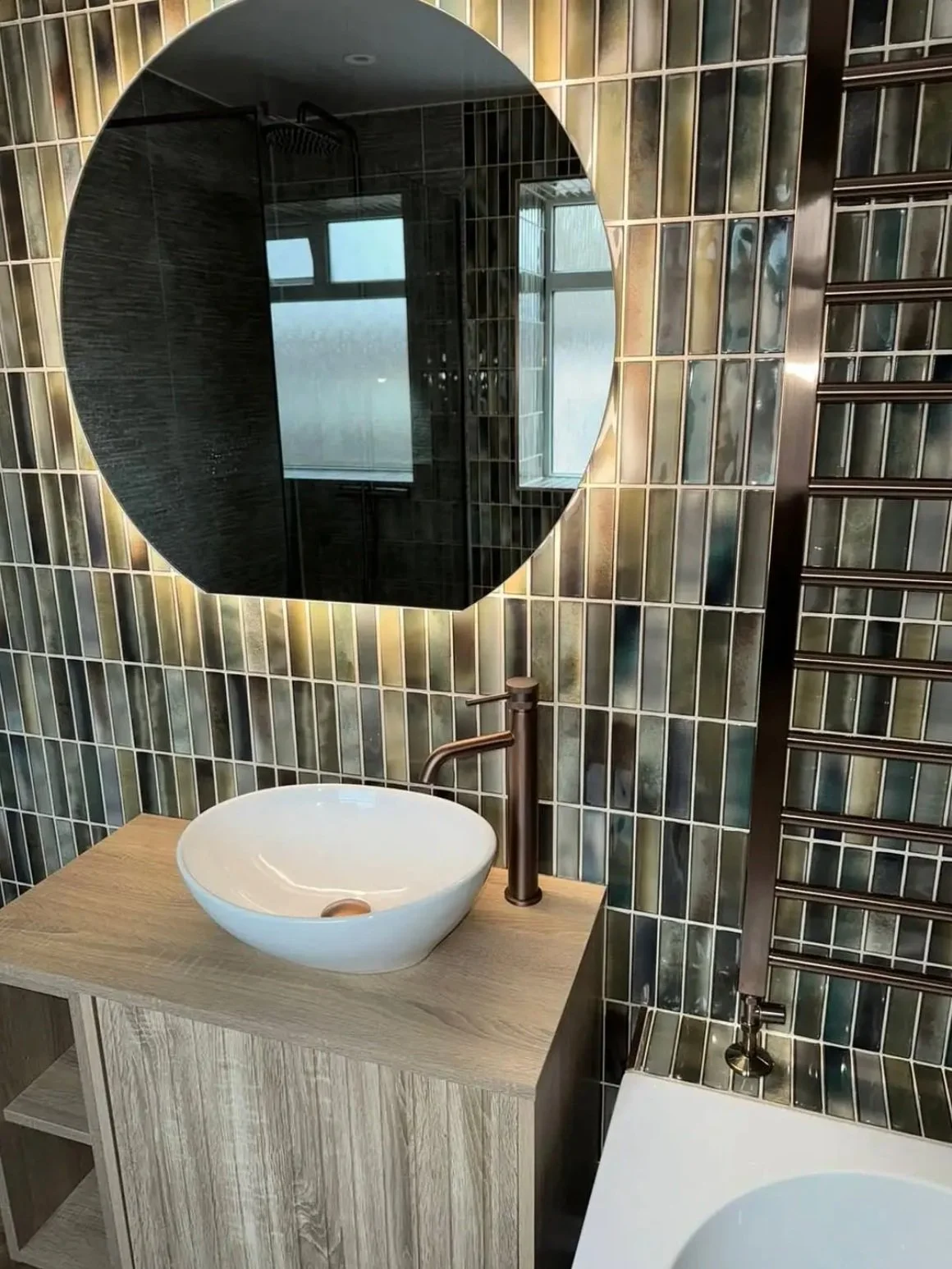 Modern bathroom in Merseyside featuring a round vessel sink on a light wood vanity, brushed bronze tap, illuminated curved mirror, and vertical multi-toned glass tiles with a contemporary towel radiator.