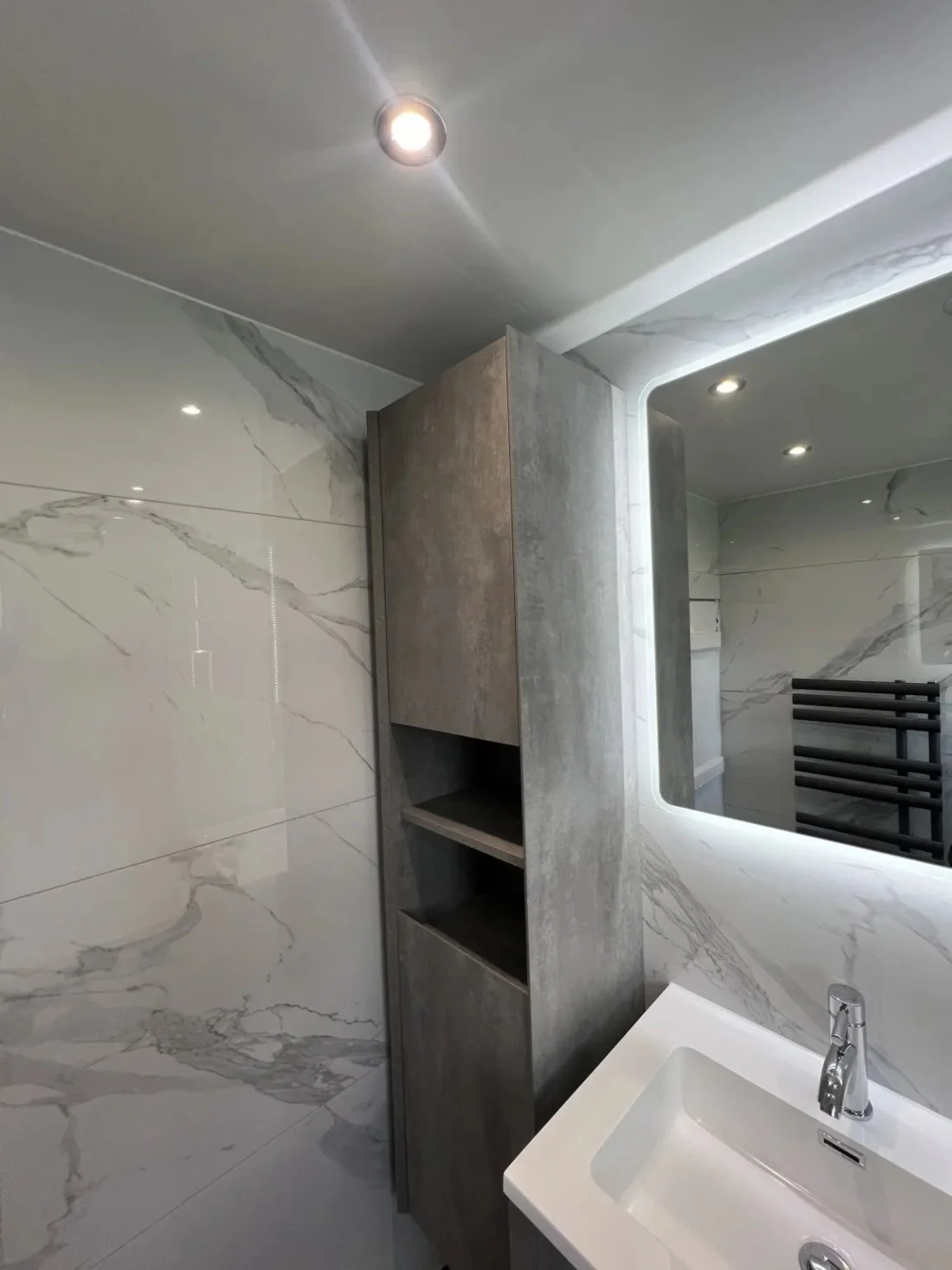 Contemporary bathroom with wall hung vanity unit, tall storage cabinet and illuminated mirror against marble effect wall panels in a Halewood home