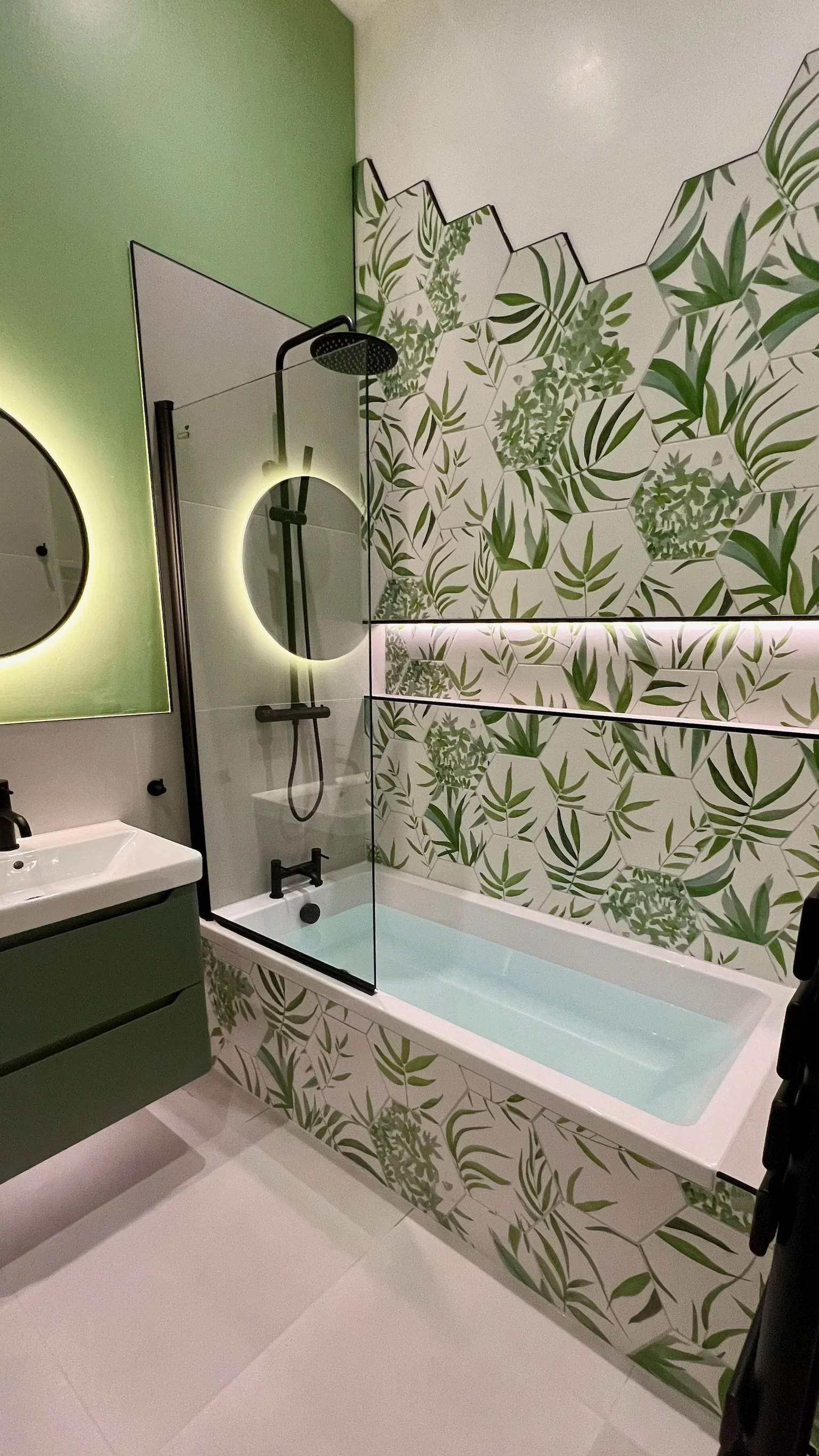 A fresh and modern bathroom renovation in Merseyside featuring a bath with overhead shower and clear glass screen. The space is brought to life by a vibrant botanical feature wall with green leaf patterns, complemented by soft green tones, a sleek va
