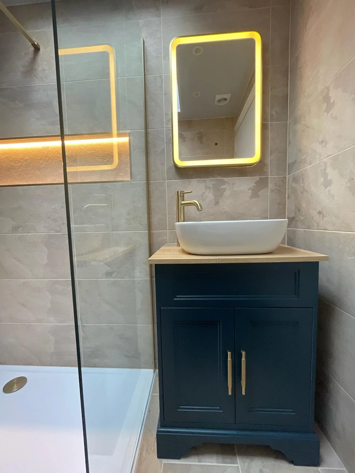 A stylish bathroom renovation in Merseyside featuring a deep blue vanity unit with a countertop basin and elegant gold fittings. The space is enhanced by a softly illuminated mirror with warm LED lighting, complemented by neutral stone-effect wall ti