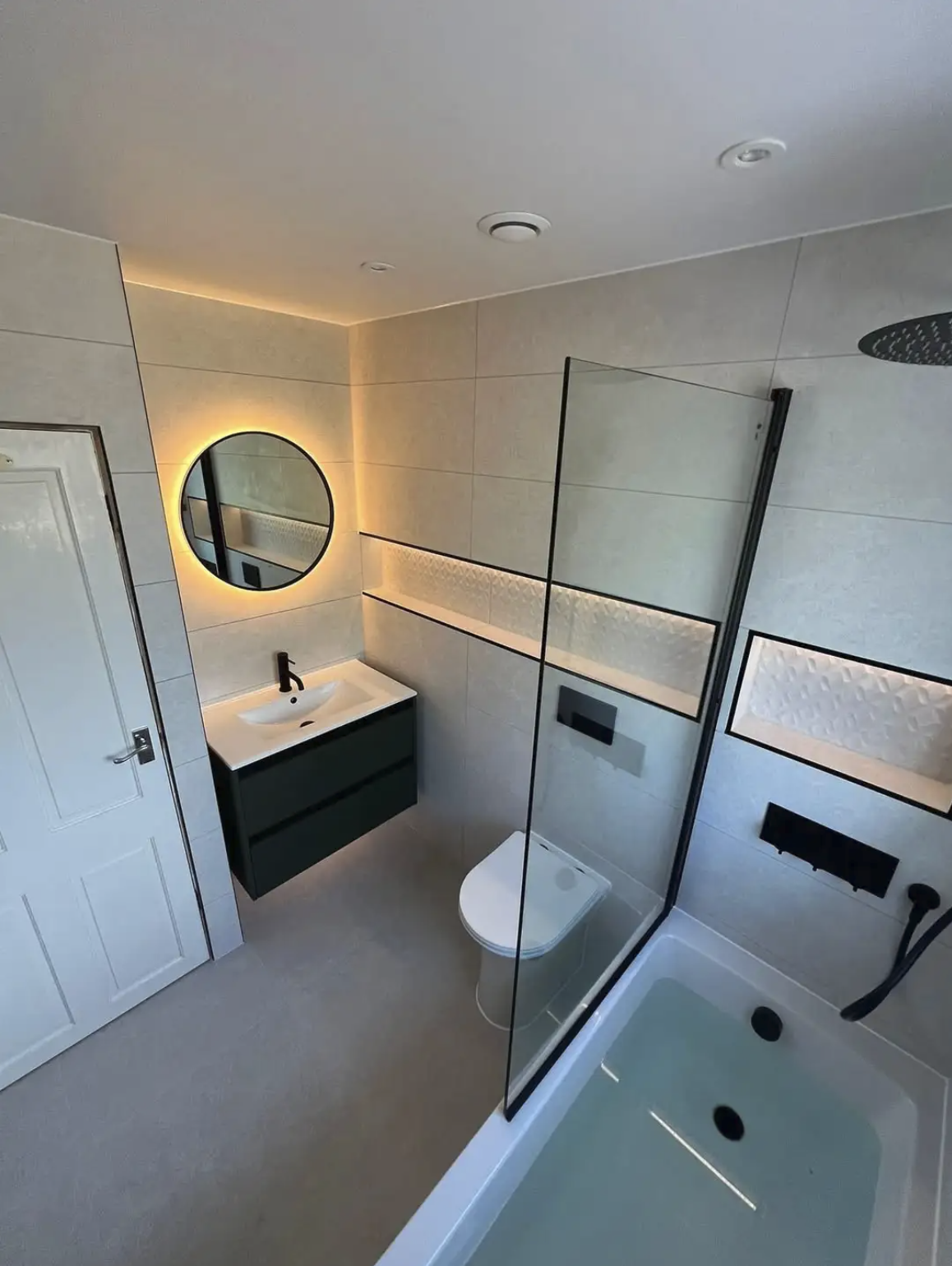 A sleek and contemporary bathroom renovation in Merseyside featuring a bath with overhead shower and a frameless glass screen. The space includes a compact wall-mounted vanity unit with an integrated basin, a round backlit mirror, and matte black fix