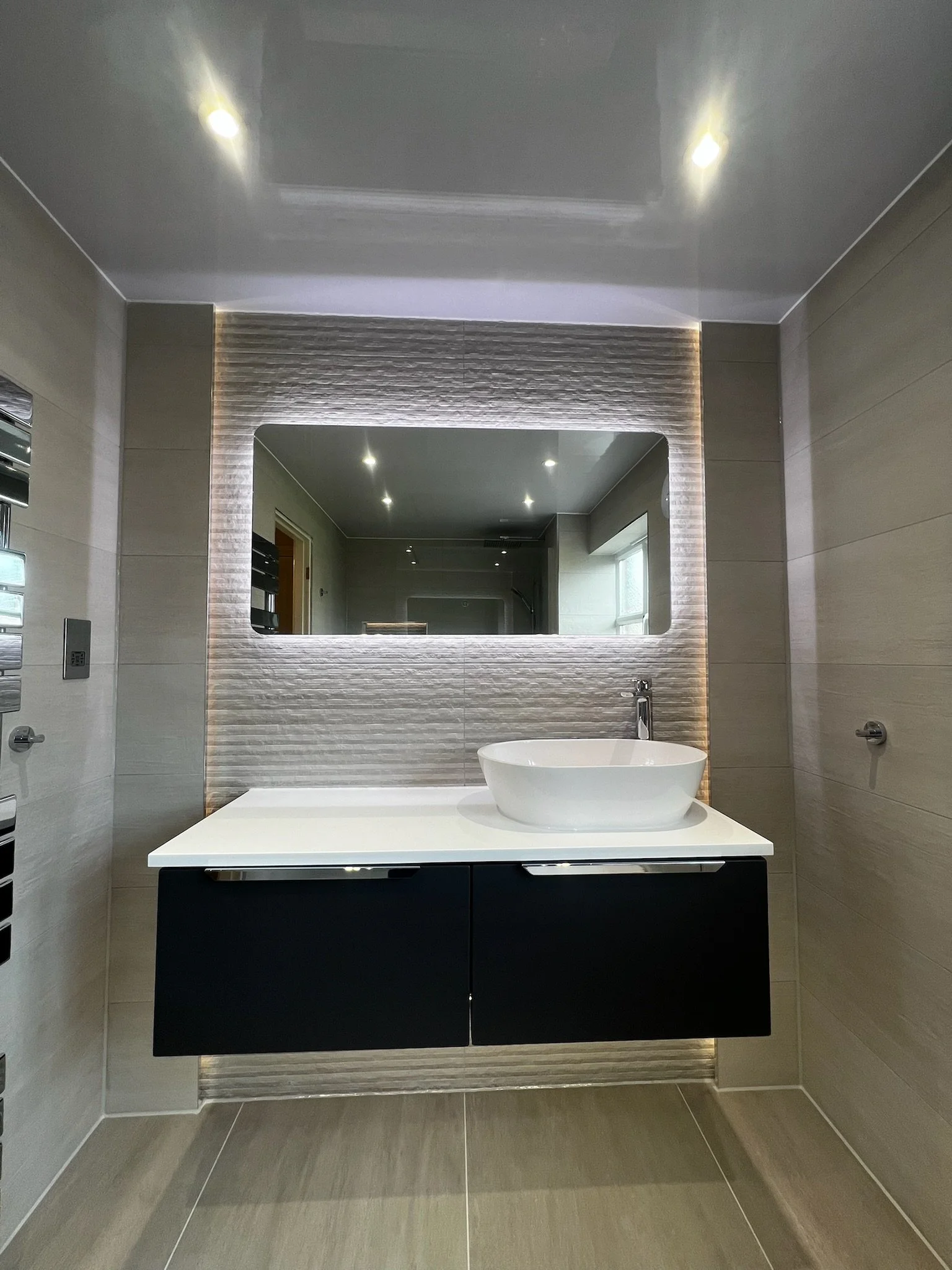 A sleek and contemporary bathroom renovation in Merseyside featuring a floating double vanity unit with a stylish countertop basin. The space is enhanced by a large illuminated mirror set within a textured feature wall, complemented by neutral tiles 