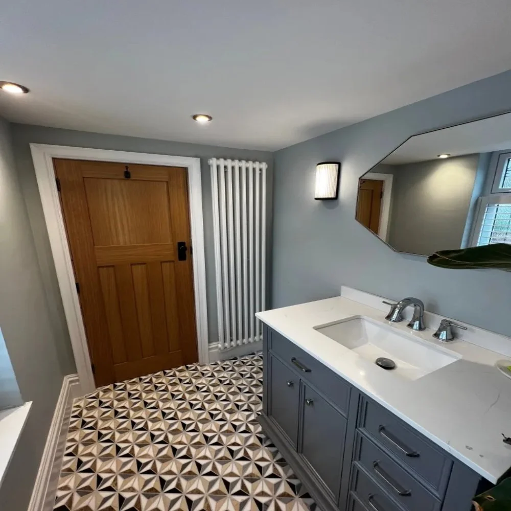 Modern downstairs toilet room in Allerton featuring a grey vanity unit with white basin, chrome tap, geometric patterned floor tiles, wall lighting, octagonal mirror, wooden door, and vertical column radiator