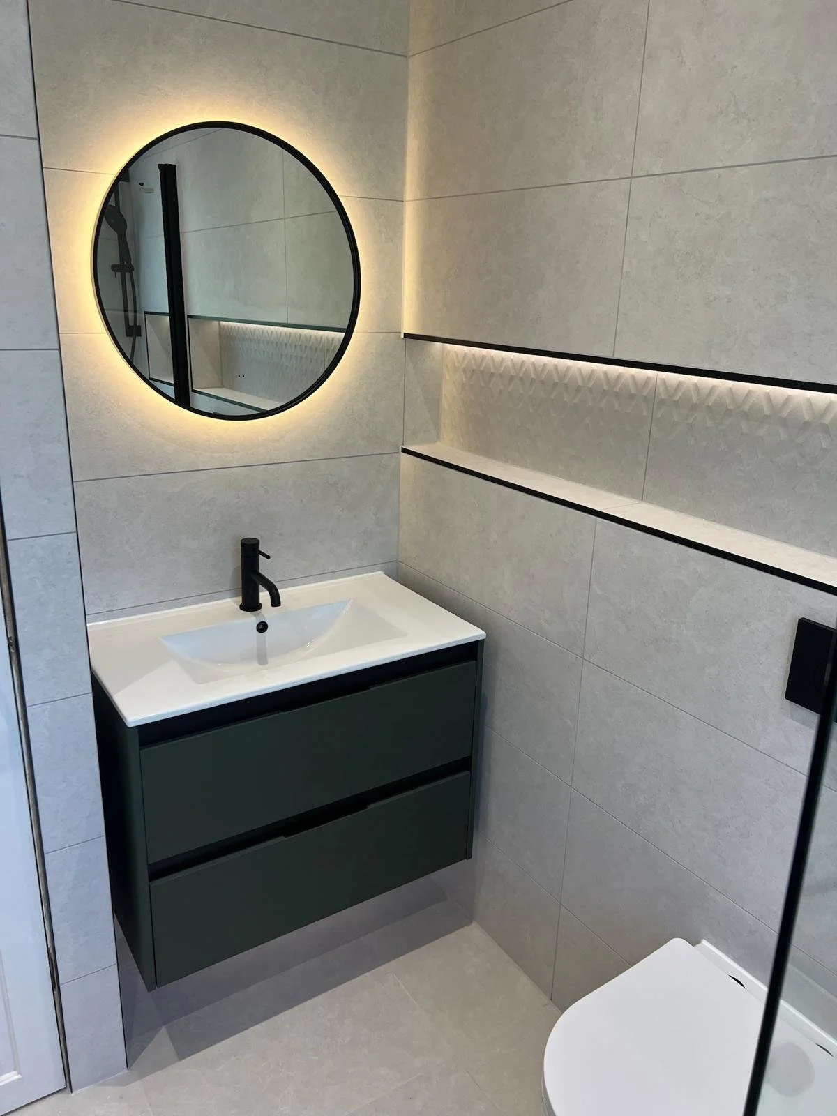 A sleek and contemporary bathroom renovation in Merseyside featuring a wall-mounted vanity unit with an integrated basin and matte black tap. The design is enhanced by a round backlit mirror, subtle recessed shelving, and large neutral tiles, creatin