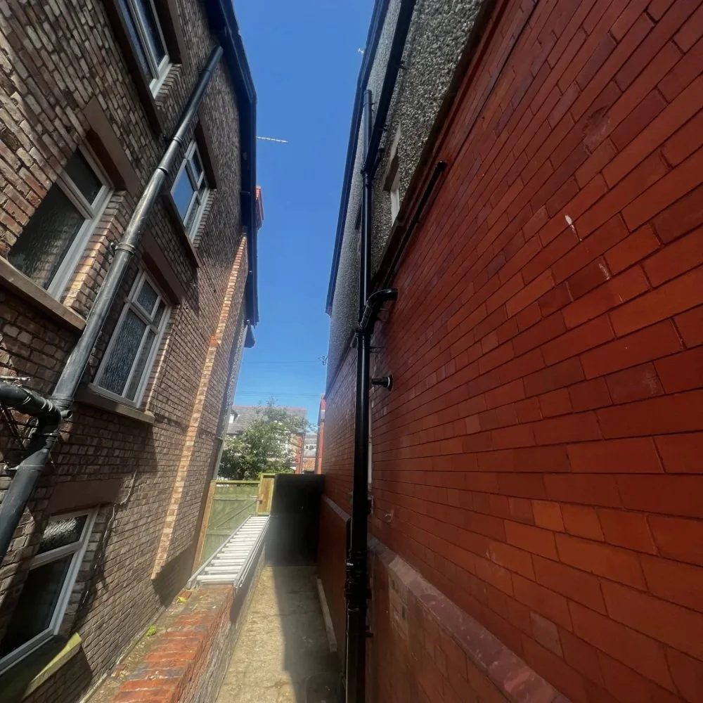 Narrow side passage between two Bebington houses with brick walls, external pipework, drainage systems, and clear blue sky above, providing access to the rear of the property
