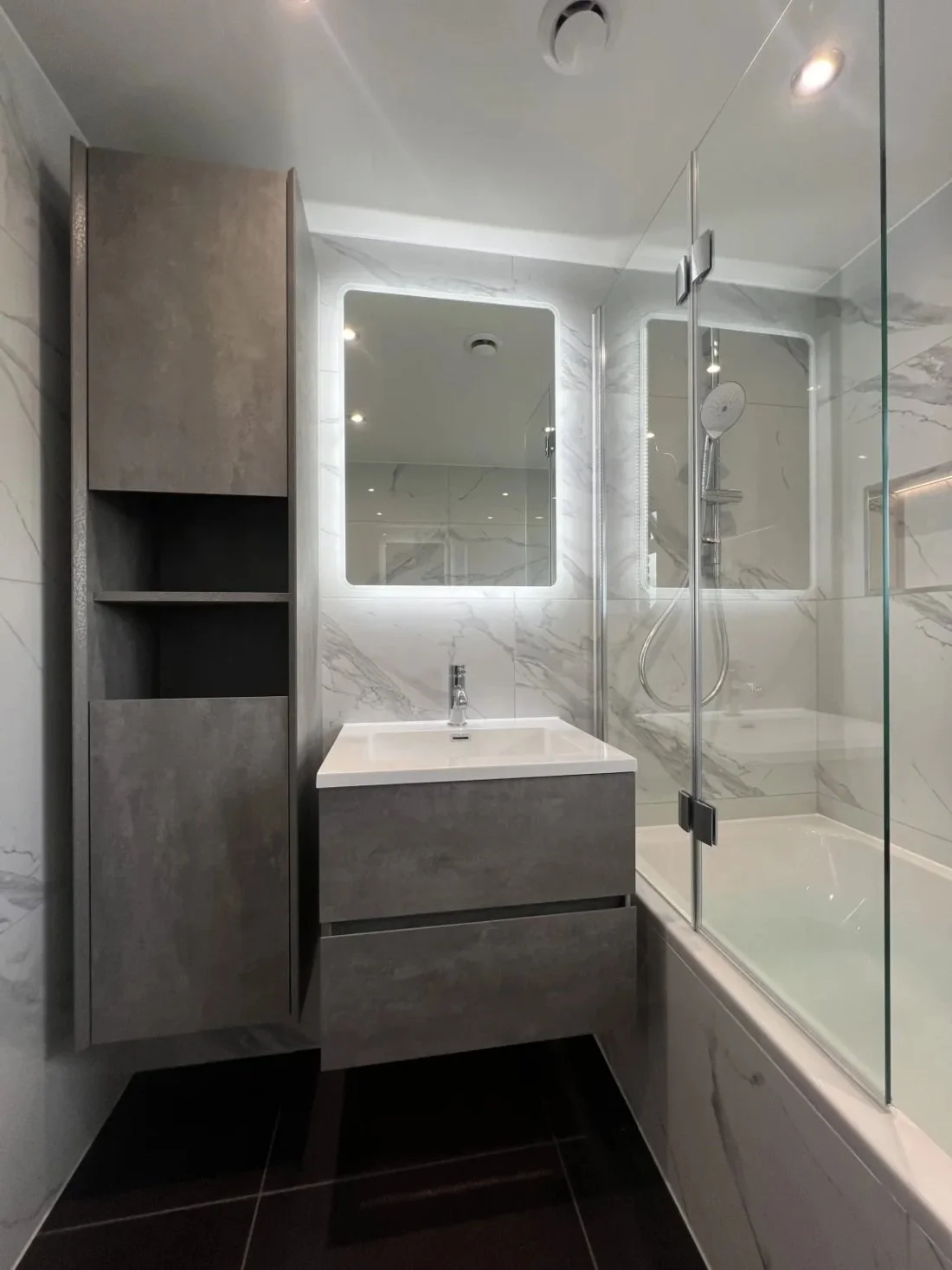 Fully renovated bathroom with wall hung vanity unit, tall storage cabinet, LED mirror, bath with glass shower screen and marble effect wall panels in a Halewood home