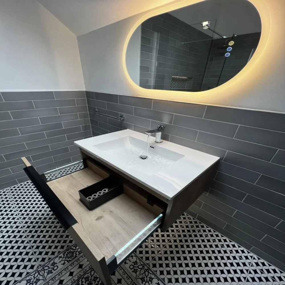Modern bathroom vanity with open drawer, integrated sink, running tap, grey wall tiles, patterned floor, and illuminated LED mirror in a Bebington renovation