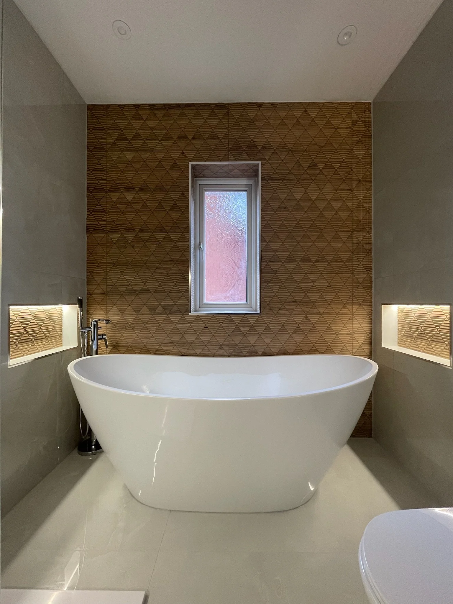 A luxurious bathroom renovation in Merseyside featuring a sleek freestanding bath set against a warm textured wood-effect feature wall. The space is enhanced by recessed alcove lighting on either side, soft neutral tones, and a central window bringin