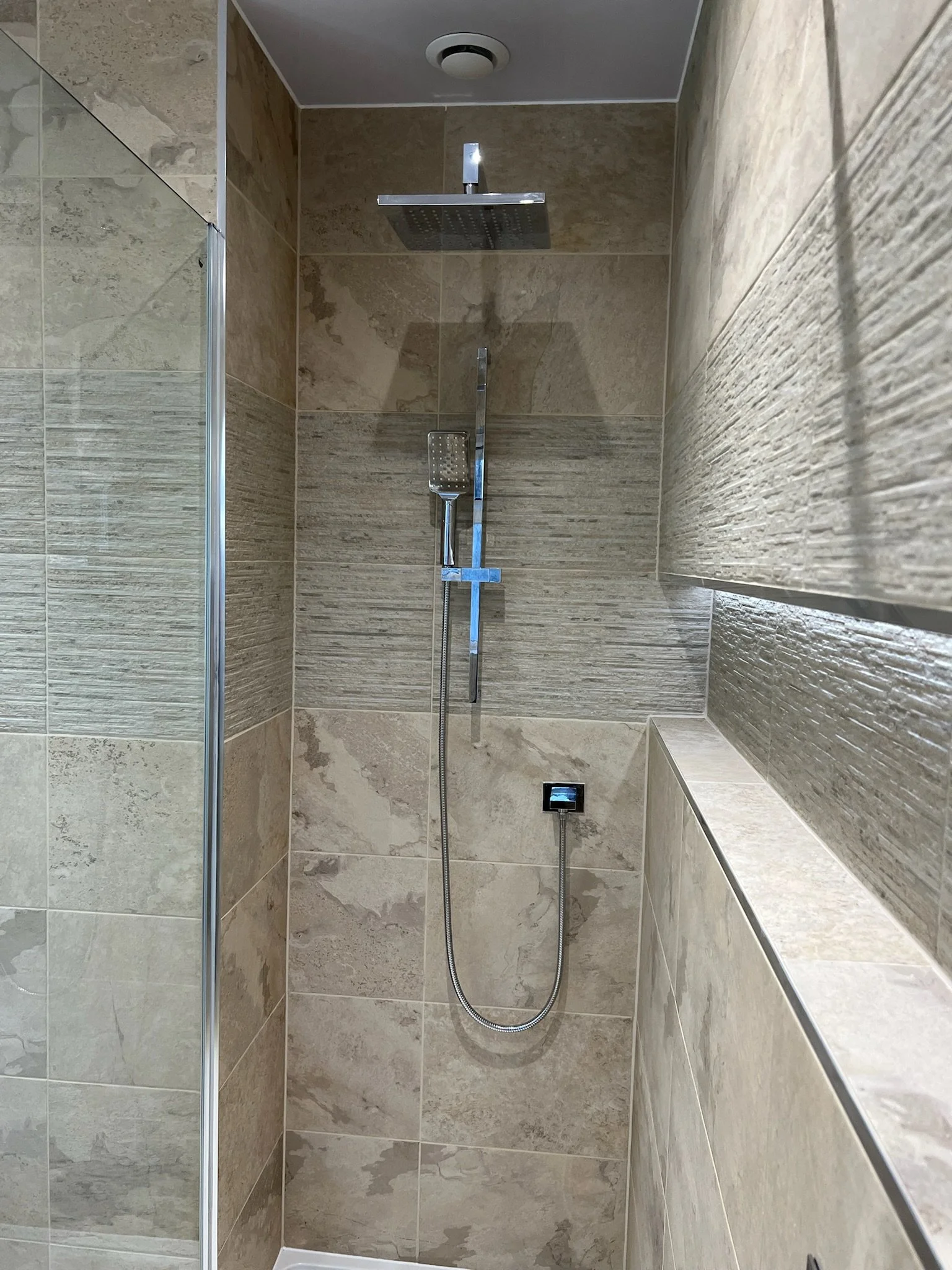 A modern walk-in shower installation in Merseyside featuring warm stone-effect wall tiles and a sleek rainfall showerhead with chrome fittings. The design includes a built-in recessed shelf for storage and a frameless glass screen, creating a clean, 