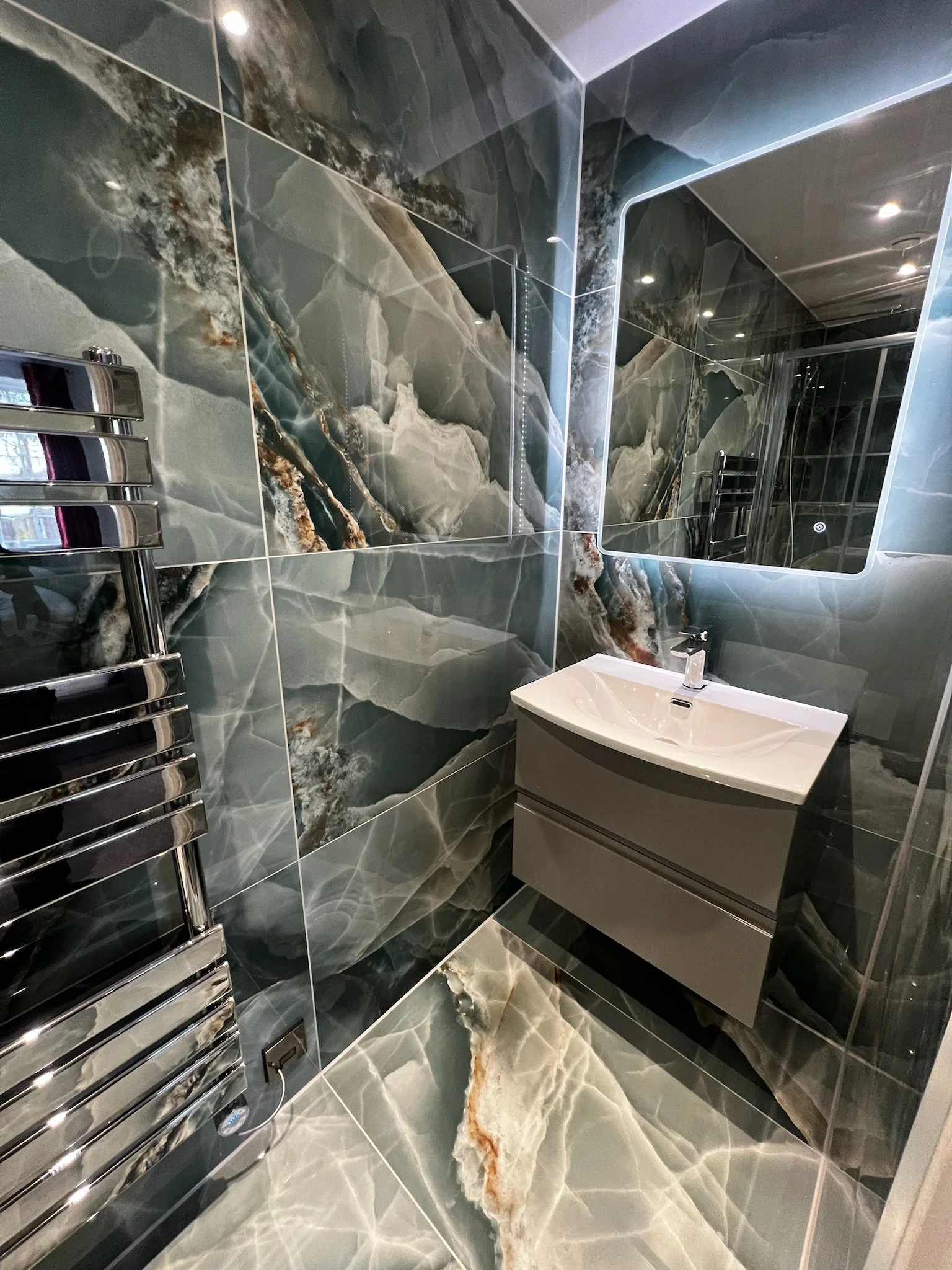 Luxury bathroom in Merseyside featuring dramatic marble-effect wall and floor tiles, wall-mounted vanity unit, illuminated mirror, chrome towel radiator, and sleek modern fittings.