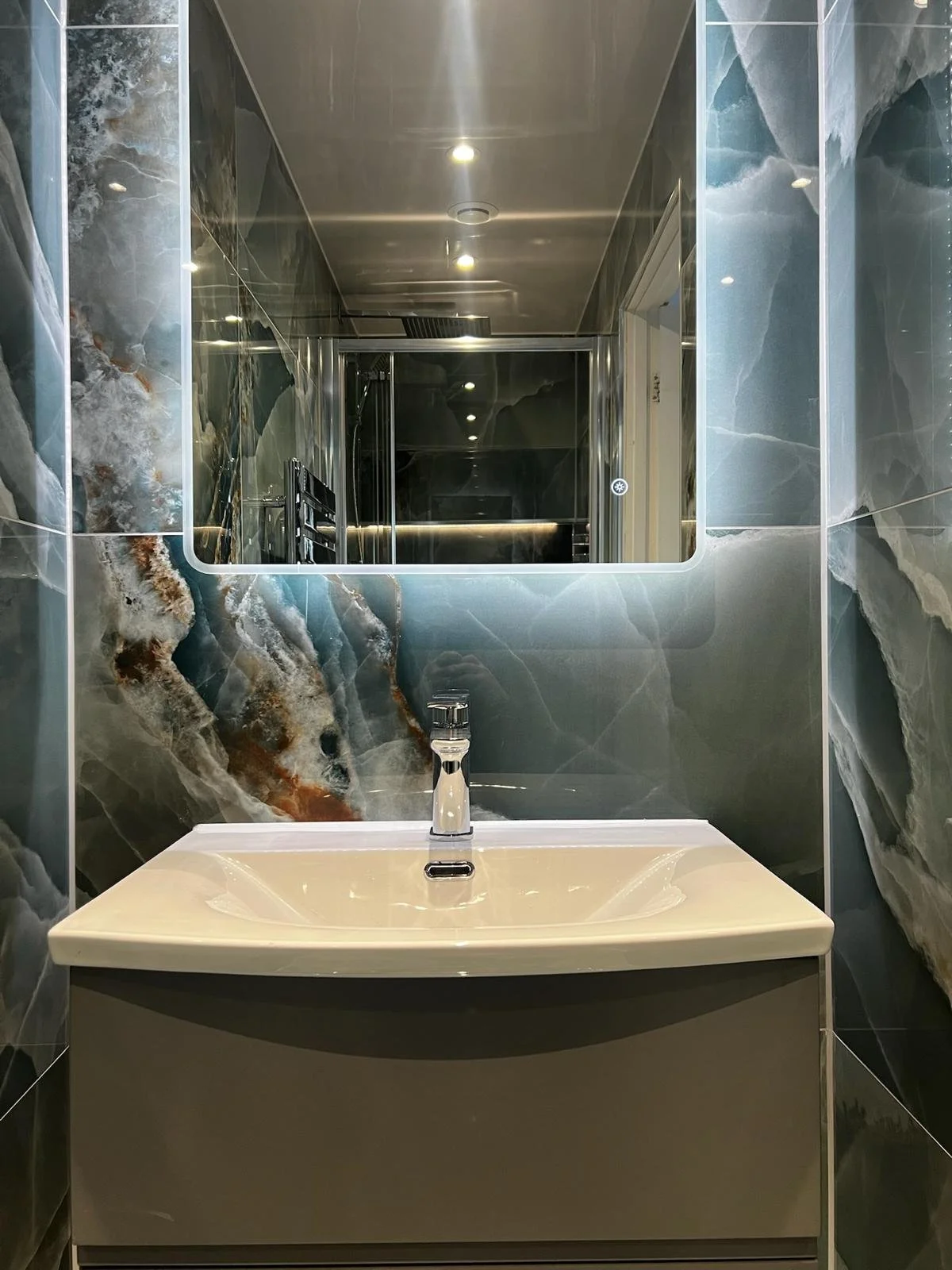 A bold and luxurious bathroom renovation in Merseyside featuring a curved vanity unit with an integrated basin, set against striking blue marble-effect wall panels. The space is enhanced by a large backlit mirror and reflective surfaces, creating a d