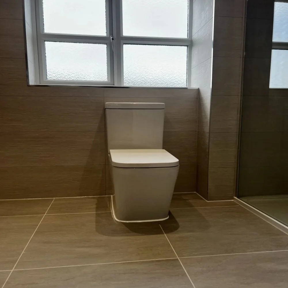 Contemporary bathroom featuring a close-coupled toilet beneath a frosted glass window, with brown wall tiles and matching floor tiles in a West Derby renovation