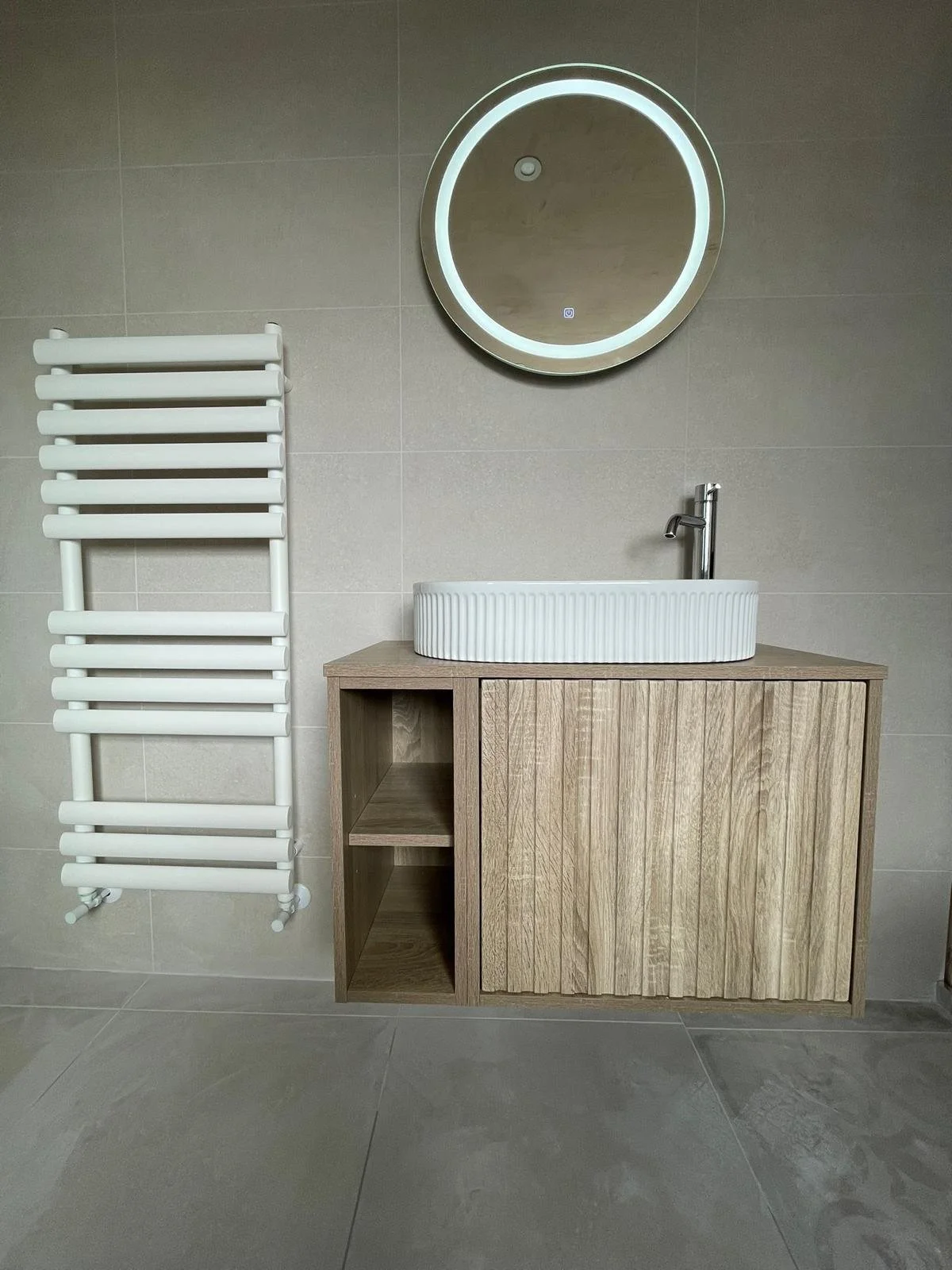 A modern bathroom installation in Merseyside featuring a natural wood vanity unit with a countertop basin and sleek chrome tap. The design is complemented by a round illuminated mirror, a white heated towel rail, and large neutral wall and floor tile