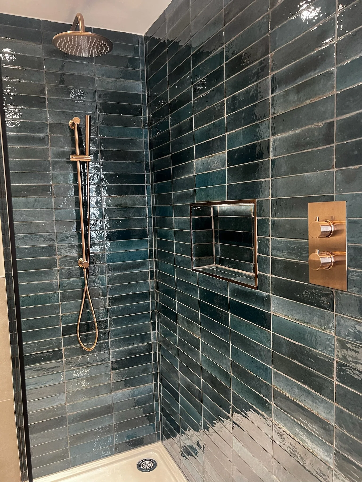 A luxurious walk-in shower installation in Merseyside featuring deep teal glossy tiles and elegant brushed gold fixtures, including a rainfall showerhead and controls. The design is enhanced by a built-in recessed niche for storage and a sleek glass 