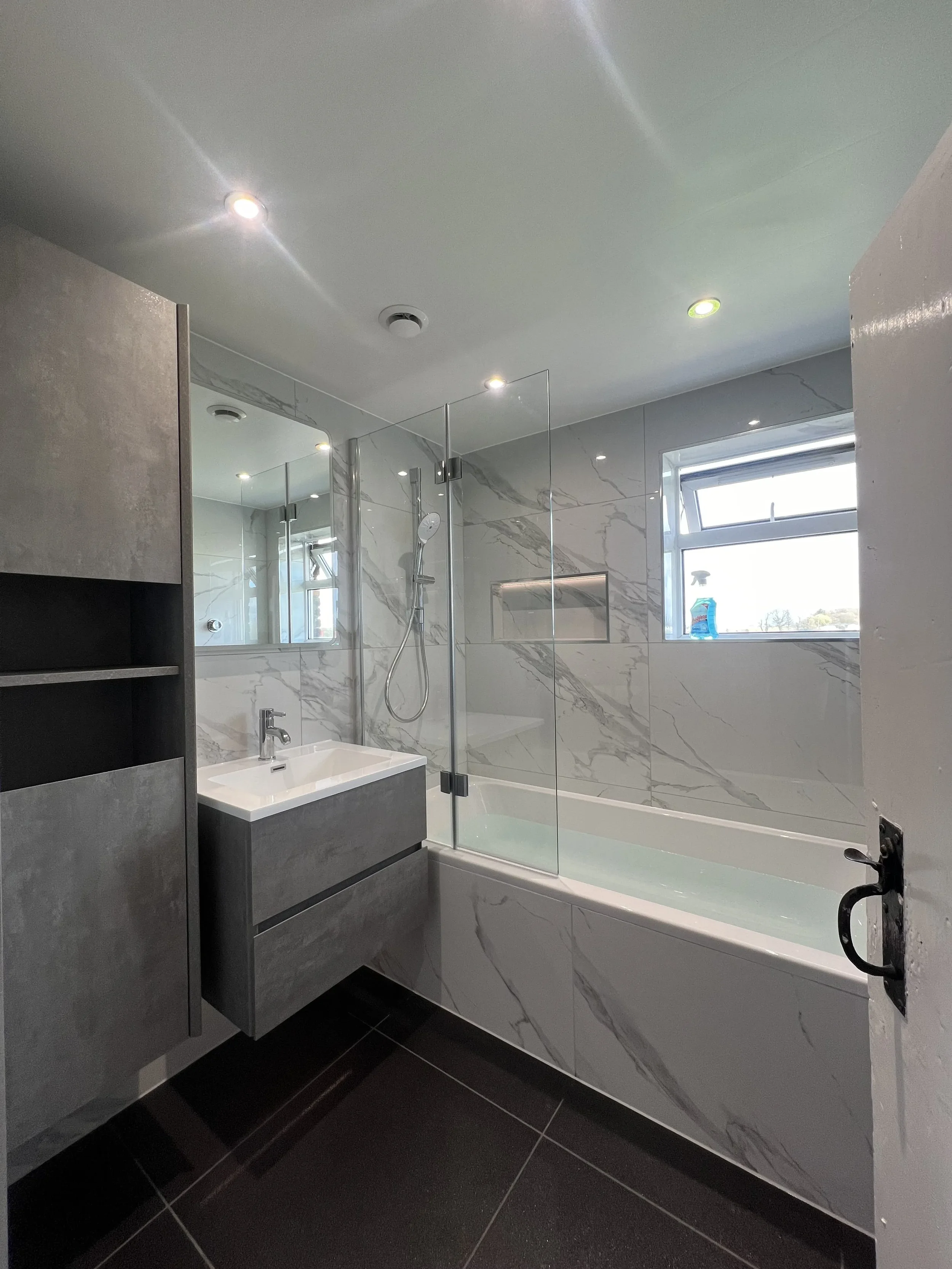 A modern bathroom renovation in Merseyside featuring a bath with overhead shower and sleek glass screen, complemented by elegant marble-effect wall panels. The space includes a wall-mounted vanity unit with an integrated basin, a large mirror, and bu