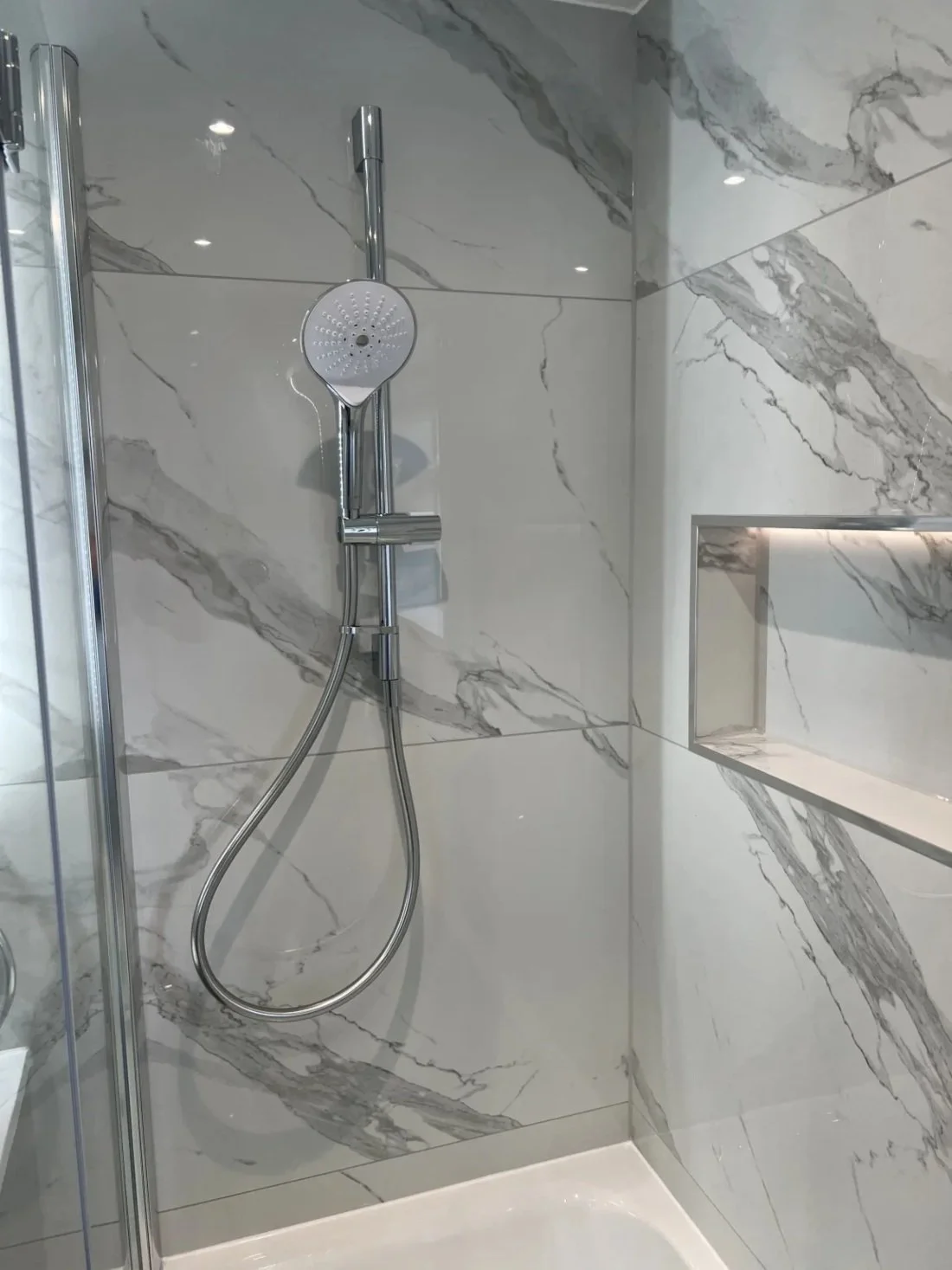 Contemporary shower over bath with glass screen, chrome shower fixture and recessed storage niche set in marble effect wall panels in a Halewood home