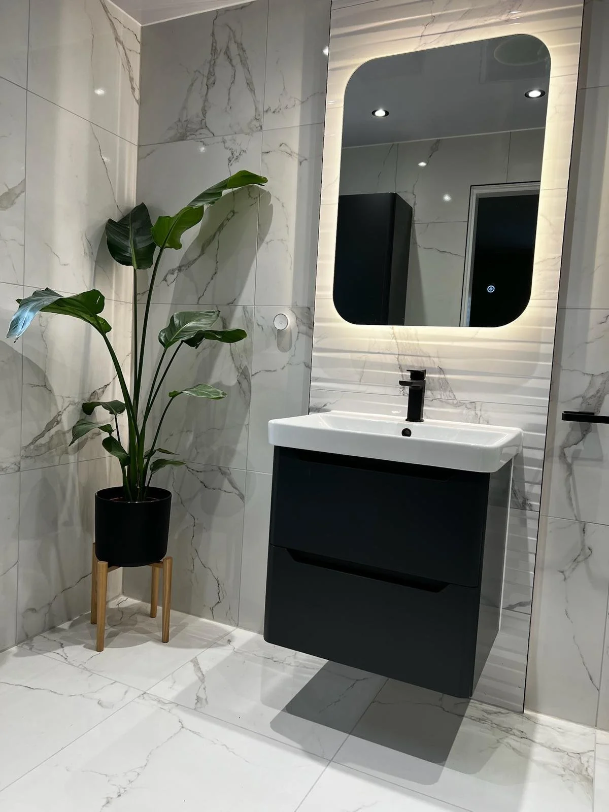 A contemporary bathroom installation in Merseyside featuring a sleek wall-mounted vanity unit with an integrated basin and matte black tap. The space is finished with elegant marble-effect wall and floor tiles, a softly illuminated mirror, and a deco