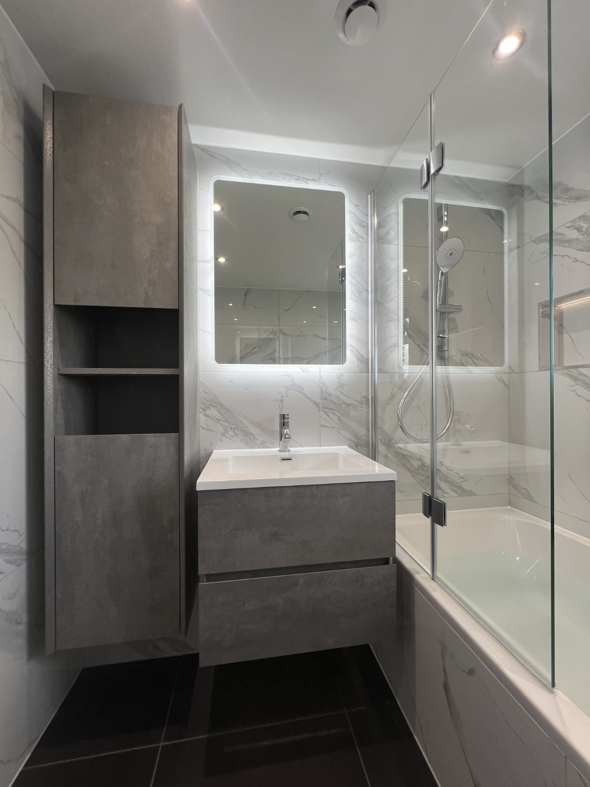 A sleek and modern bathroom renovation in Merseyside featuring a wall-mounted vanity unit with a minimalist integrated basin and a large illuminated mirror. The space includes a bath with an overhead shower and glass screen, complemented by marble-ef