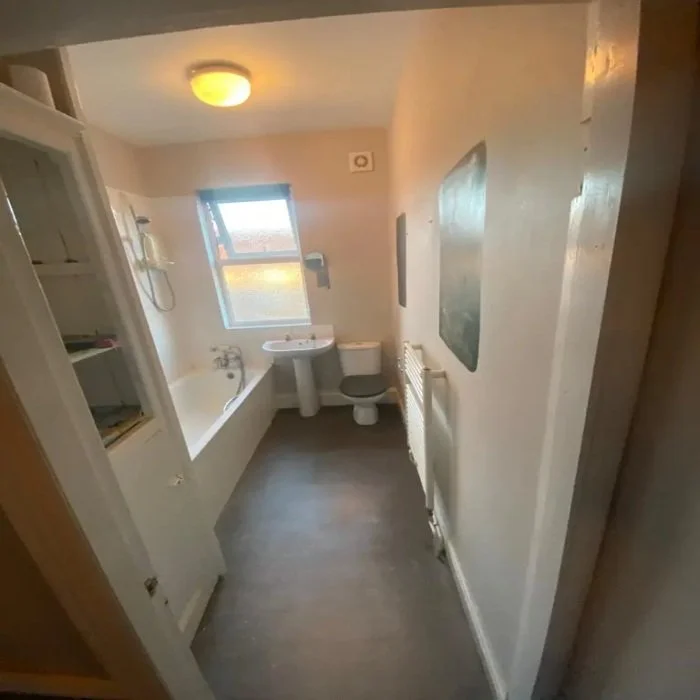 Narrow outdated bathroom in Mossley Hill featuring built-in bath with shower, pedestal sink, close-coupled toilet, radiator, and vinyl flooring before modern renovation.