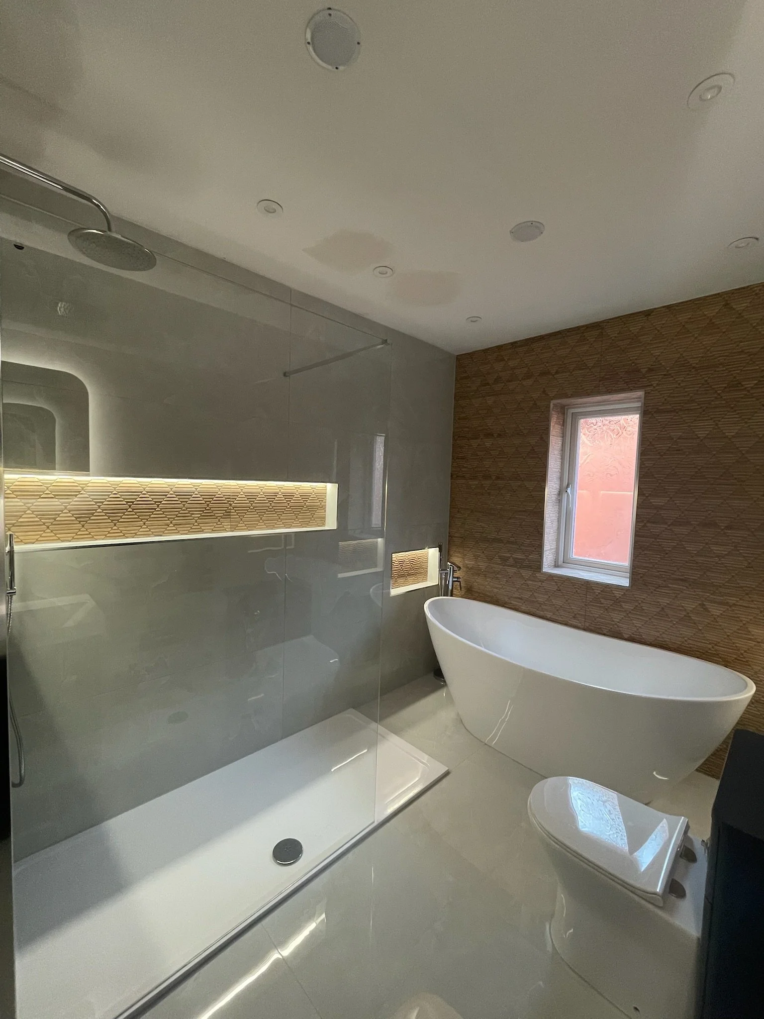 A modern and spacious bathroom renovation in Merseyside featuring a walk-in shower with sleek glass screen and soft ambient LED lighting. The design includes a freestanding bath set against a warm textured feature wall, complemented by neutral tiles 