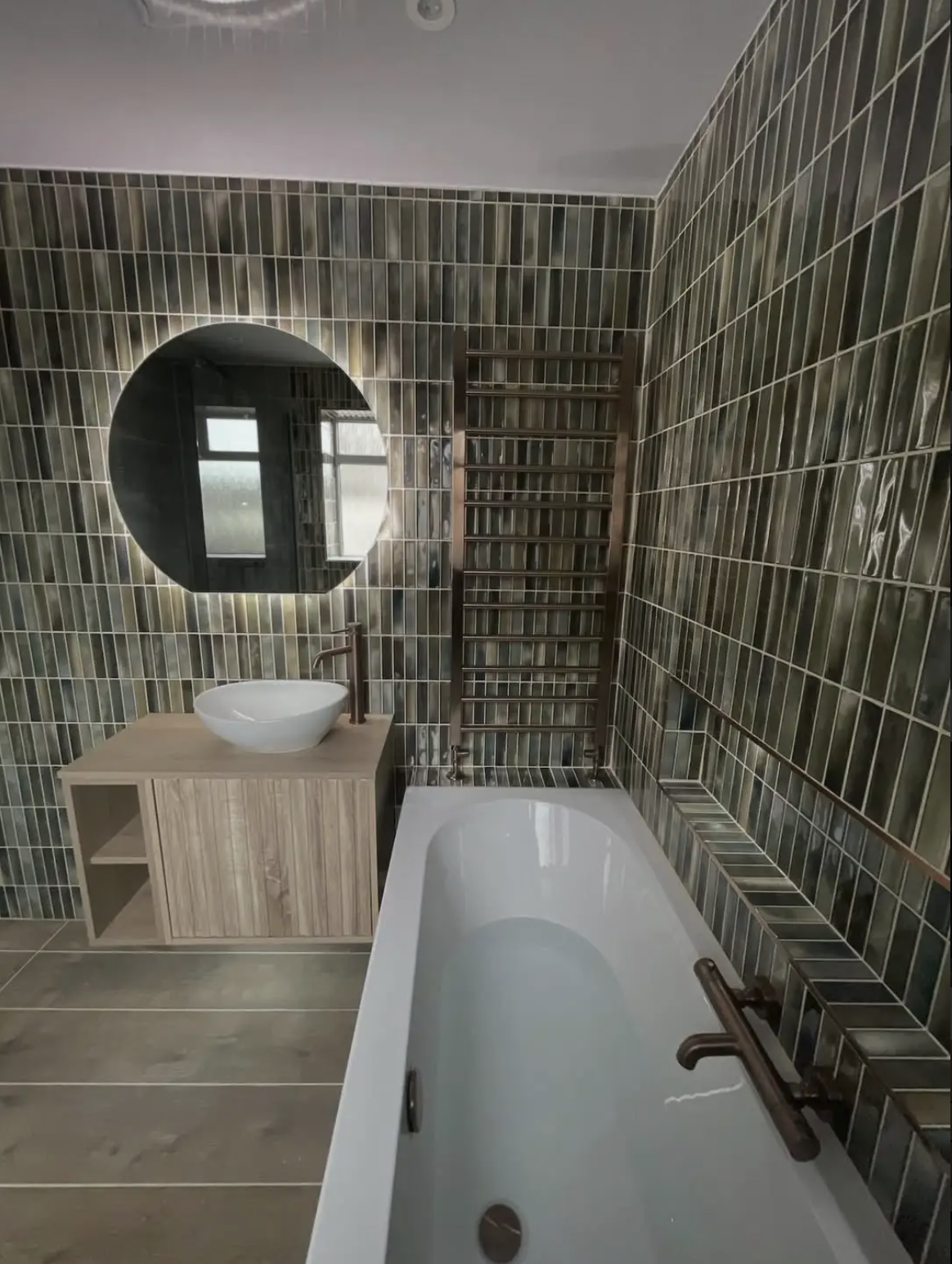 A stylish and contemporary bathroom renovation in Merseyside featuring rich green vertically stacked tiles that create a bold, textured feature throughout the space. The design includes a sleek built-in bath with bronze fixtures, a floating wood-effe