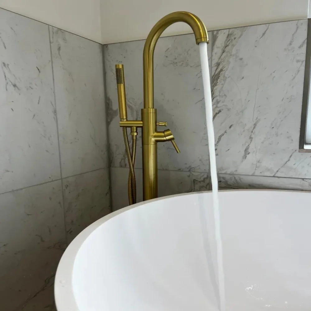 Close-up of a freestanding bath in Allerton featuring a brushed brass floor-mounted tap with handheld shower attachment, set against marble effect wall tiles with running water filling the tub