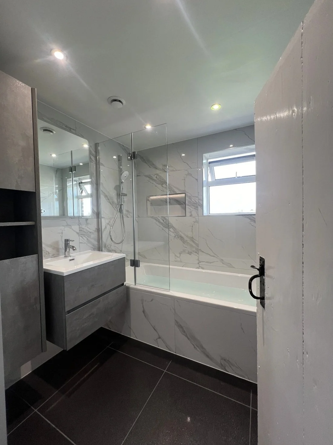 Stylish modern bathroom featuring marble effect wall panels, bath with glass shower screen, floating vanity unit and black floor tiles in a Halewood home