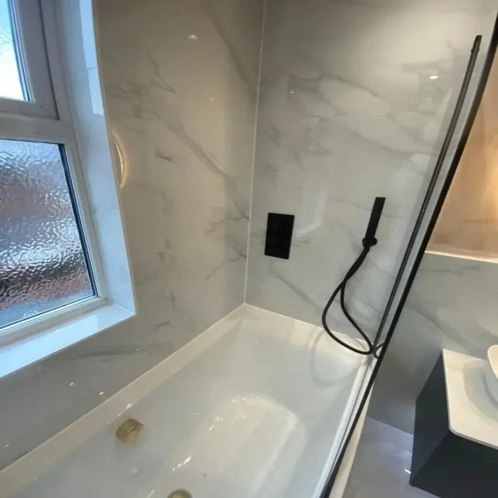 Close-up of bathroom in Mossley Hill showing marble-effect wall panels, built-in bath with gold waste, black wall-mounted shower controls and handset, glass shower screen, and frosted window.