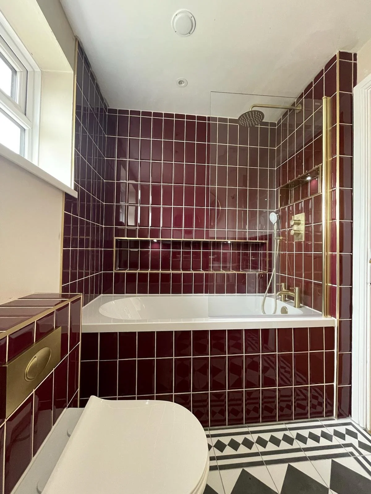 A bold and contemporary bathroom renovation in Merseyside featuring rich burgundy gloss tiles surrounding a bath with an overhead shower. The design includes a built-in recessed shelf, a wall-mounted vanity with warm wood accents, and striking geomet