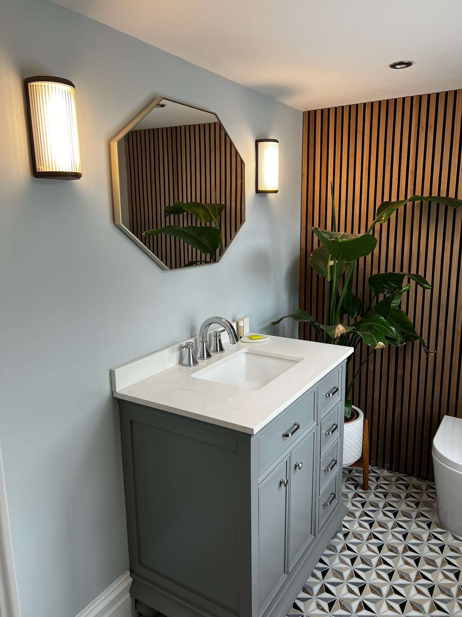 A contemporary bathroom renovation in Merseyside featuring a stylish grey vanity unit with a classic basin and chrome fittings. The space is enhanced by a unique hexagonal mirror, warm wall lighting, and a striking wood slatted feature wall, compleme